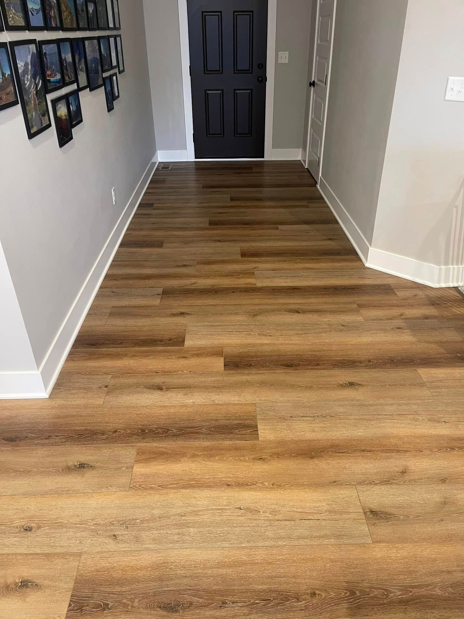 Hardwood-style flooring in a hallway, leading to a black door. White trim and grey walls are present.