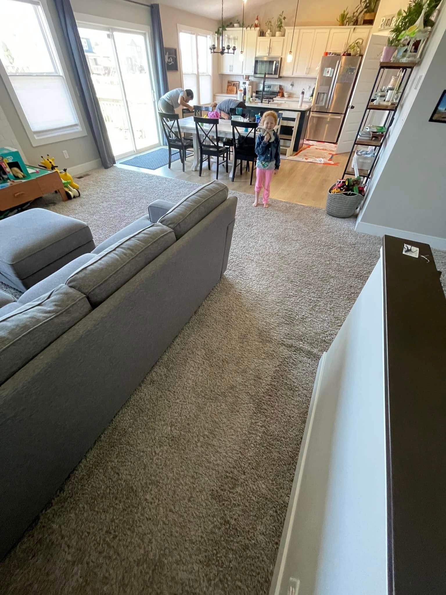 Living room with gray sofa, large rug, and person in kitchen area.