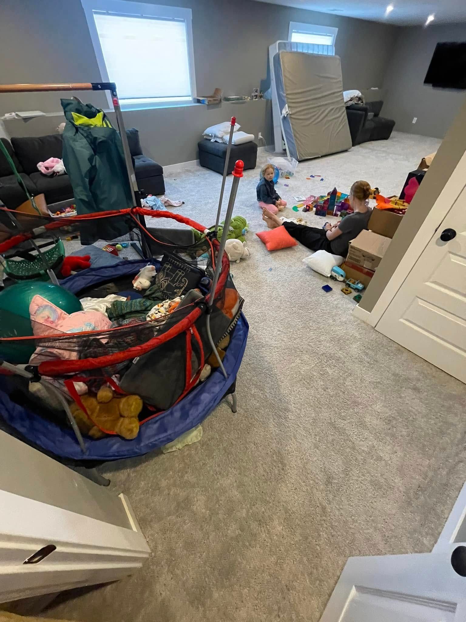 A messy playroom with a trampoline, child, and adult surrounded by toys and furniture.