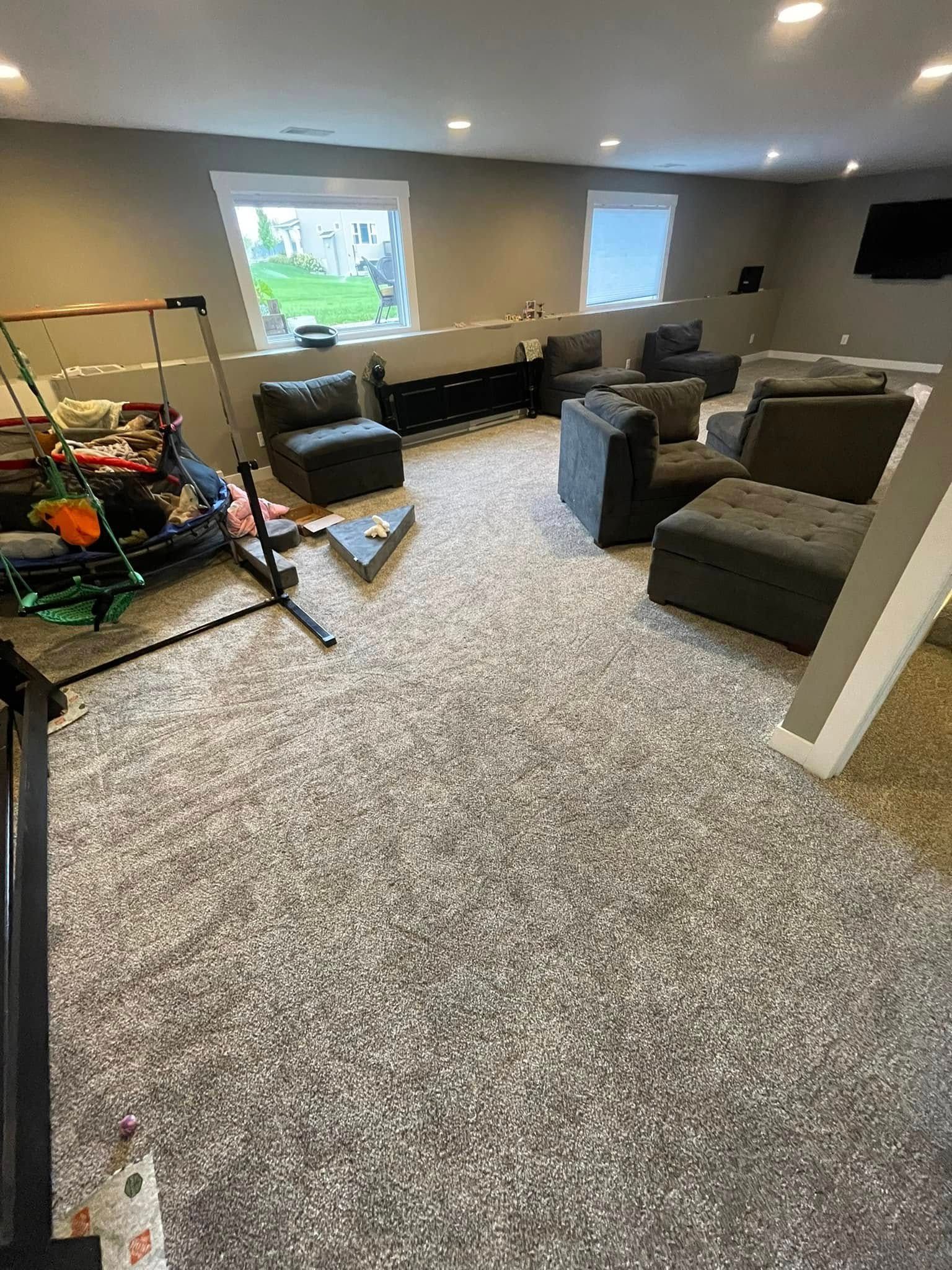 Gray carpeted basement with gray armchairs, a black entertainment console, and clutter. Windows and recessed lighting.