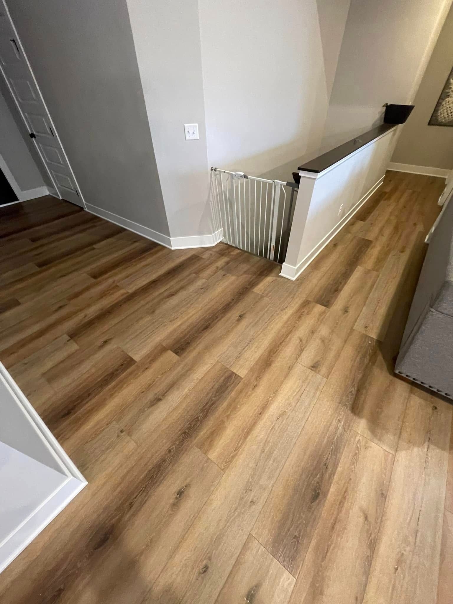 Hardwood-like flooring in room with a baby gate at top of staircase. Light brown wood, white walls.
