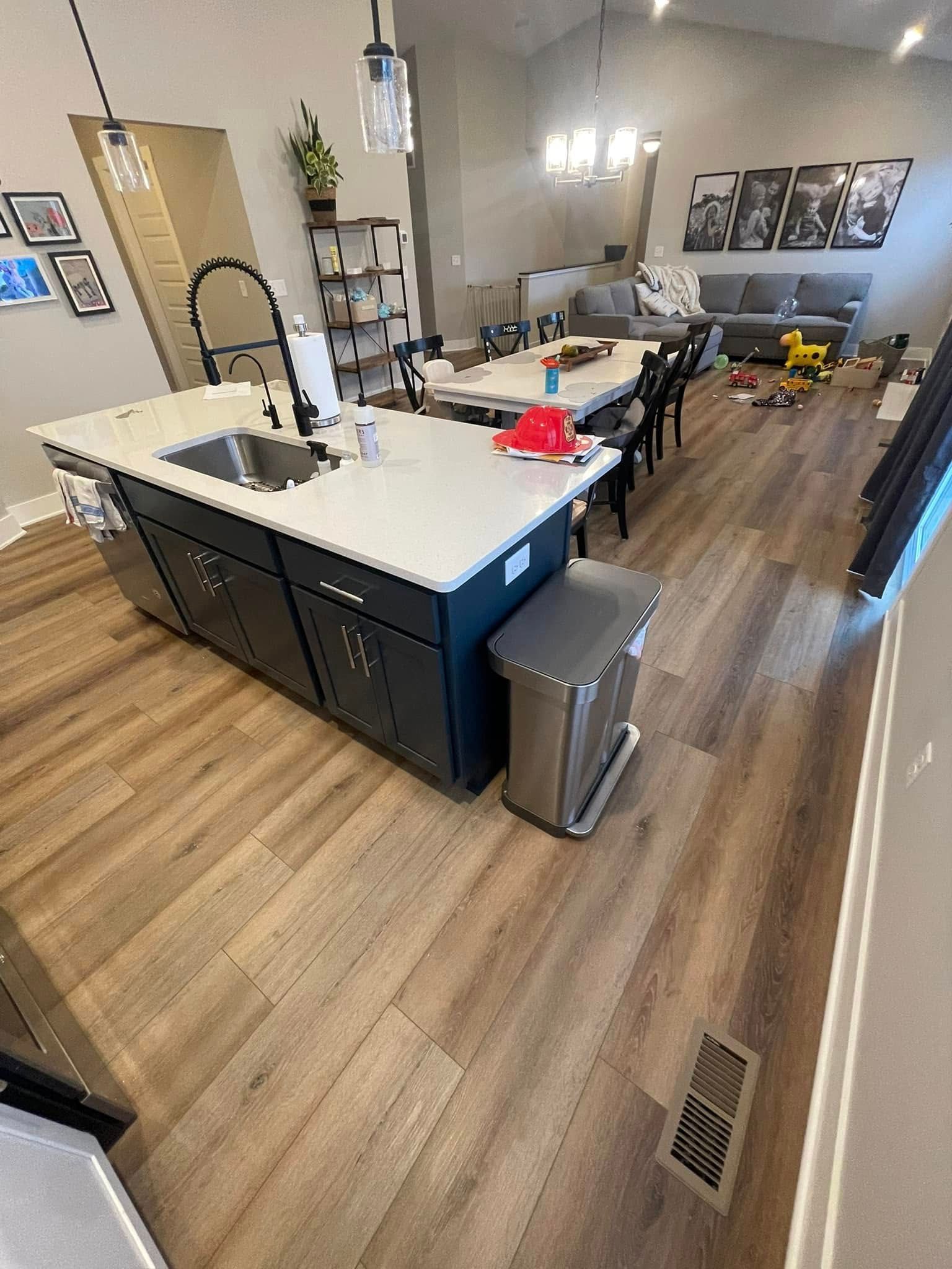 Kitchen island with sink, navy cabinets, white countertop, trash can, and wood-look floors. Dining area and living room visible.
