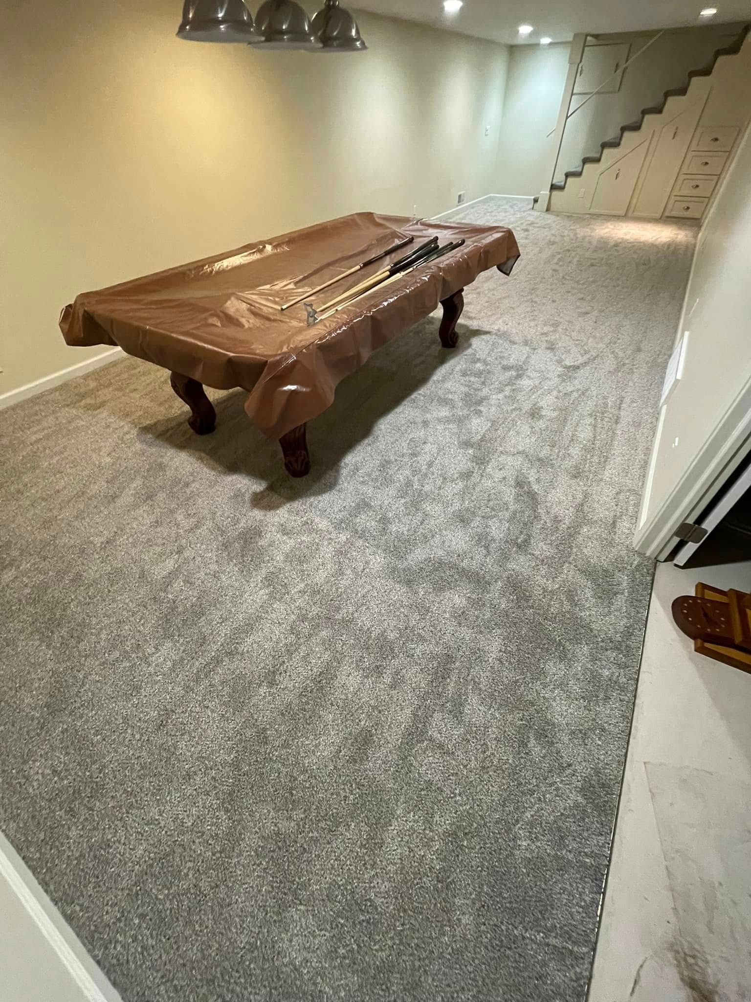 A basement recreation room with a covered pool table on grey carpet. Stairs and white walls in the background.