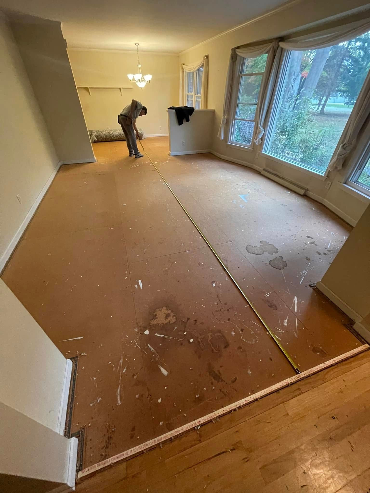 Person in protective gear sprays floor in a room with bay windows. Brown floor, tape measure, white walls, and a chandelier are visible.