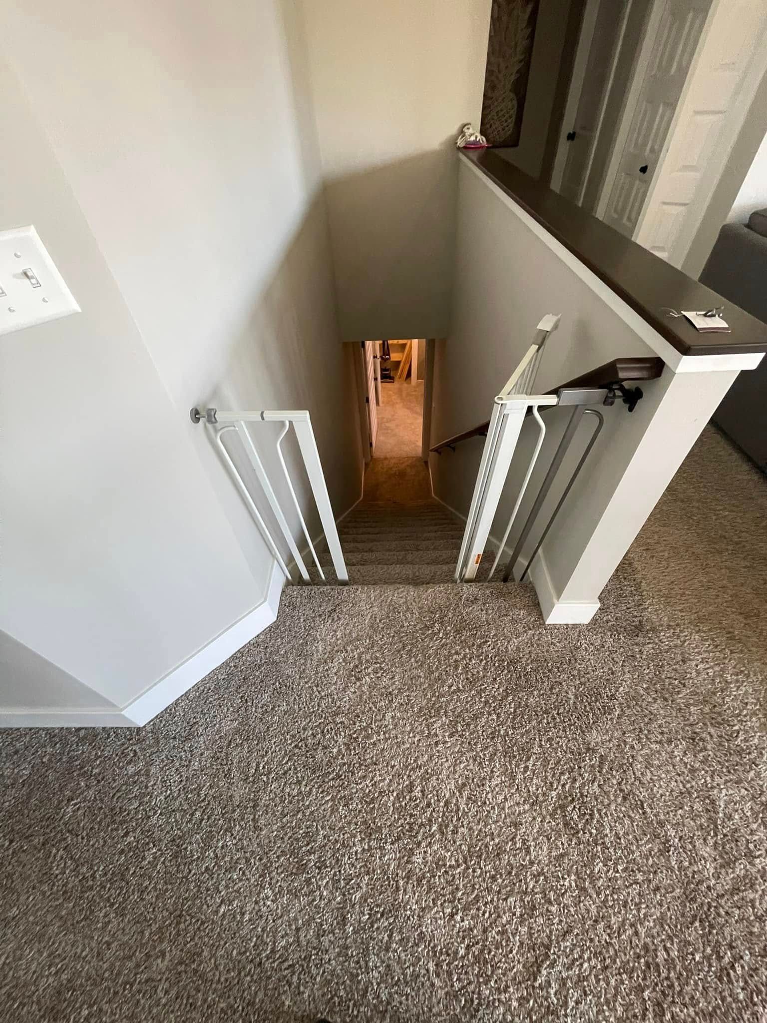 Stairway leading down, with safety gates at top. Light walls, carpeted floor, dark wooden handrail.