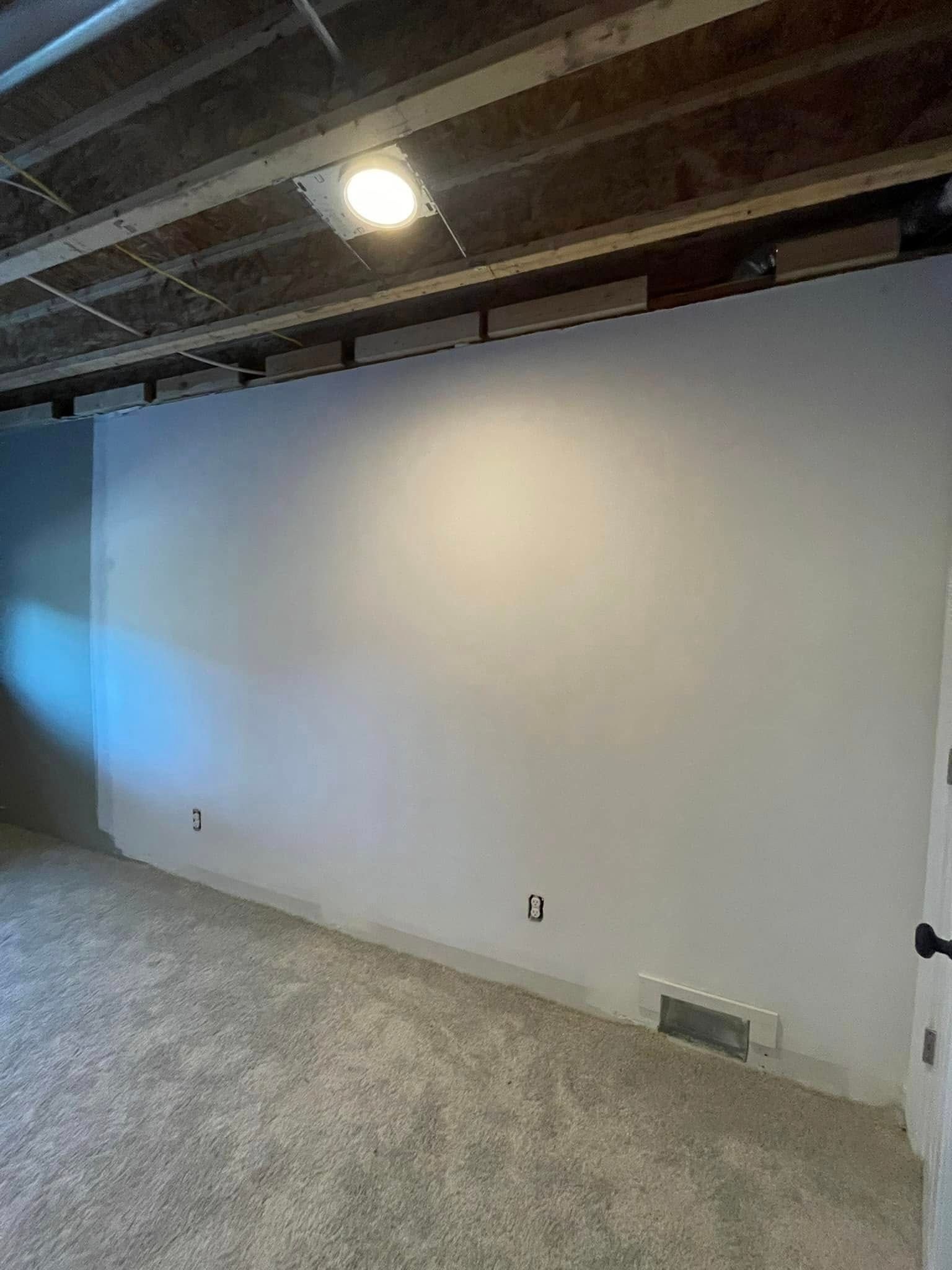 White wall in a room with gray carpet, under exposed ceiling beams. A light shines on the wall.