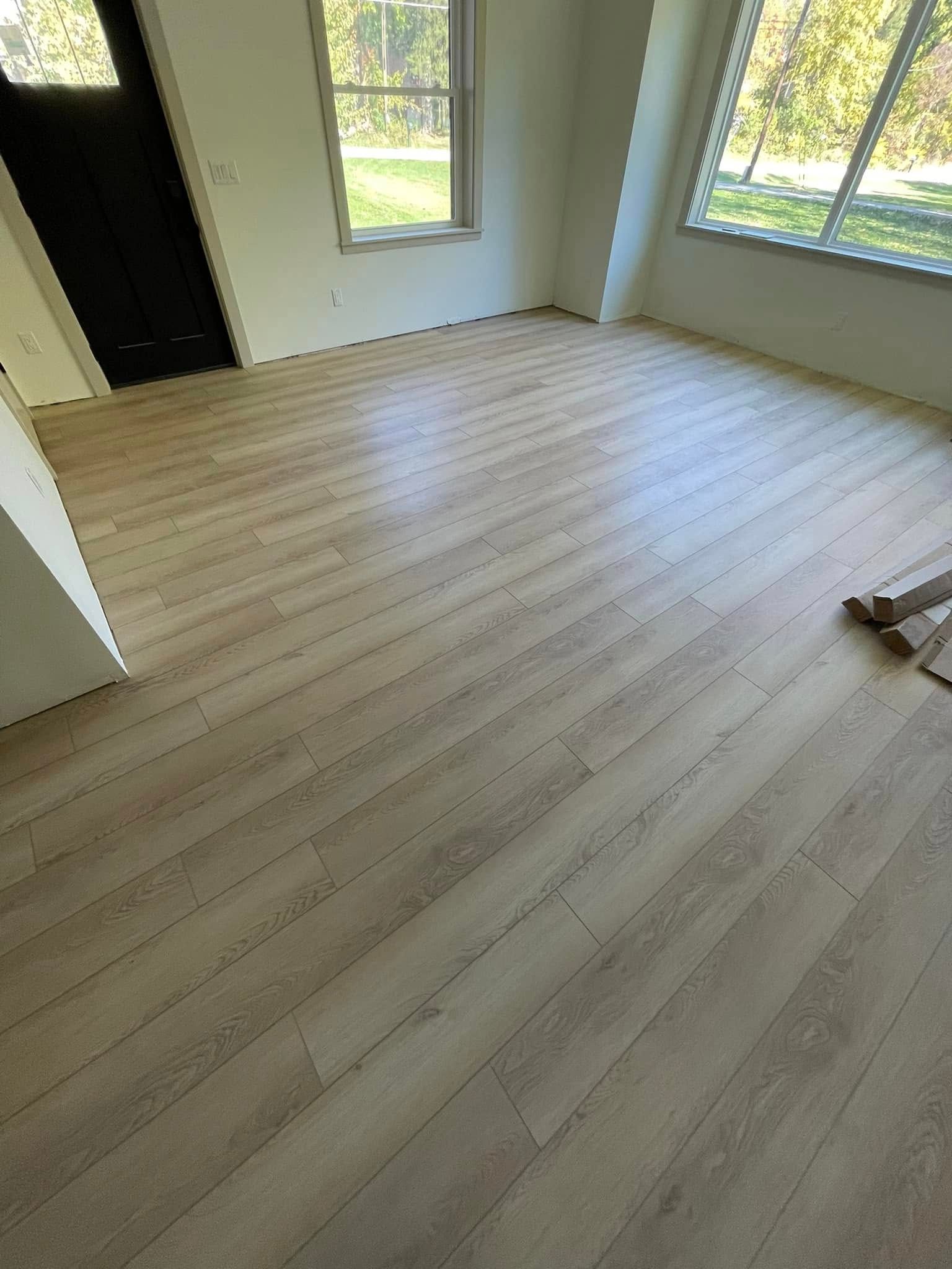 Light wood-look flooring installed in a room with windows and a dark door; unfinished walls.