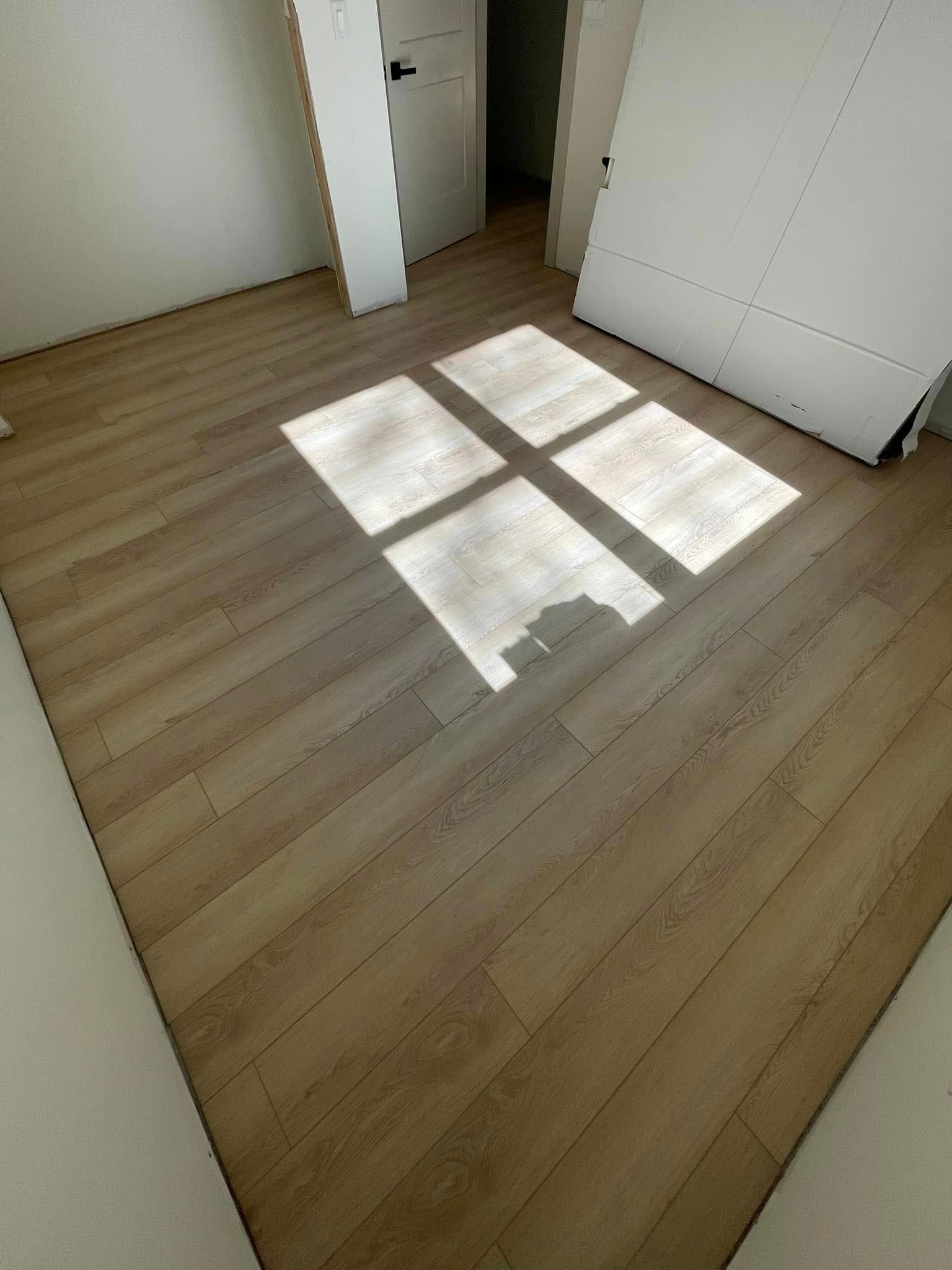 Sunlight casts a square shadow on a wooden floor in an empty room, white walls and a door visible.