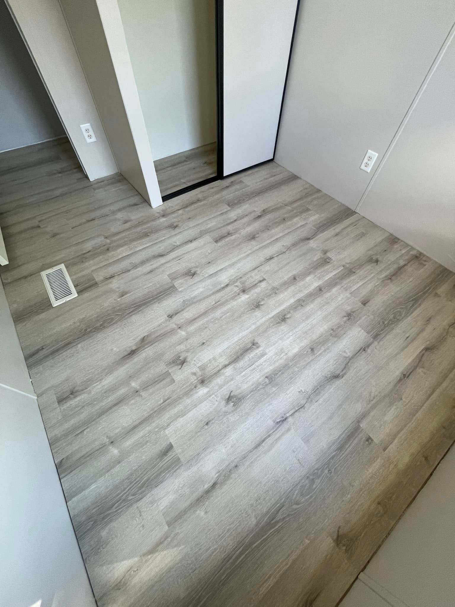 Light gray wood-look flooring in a room with white walls. Air vent and electrical outlets are visible.