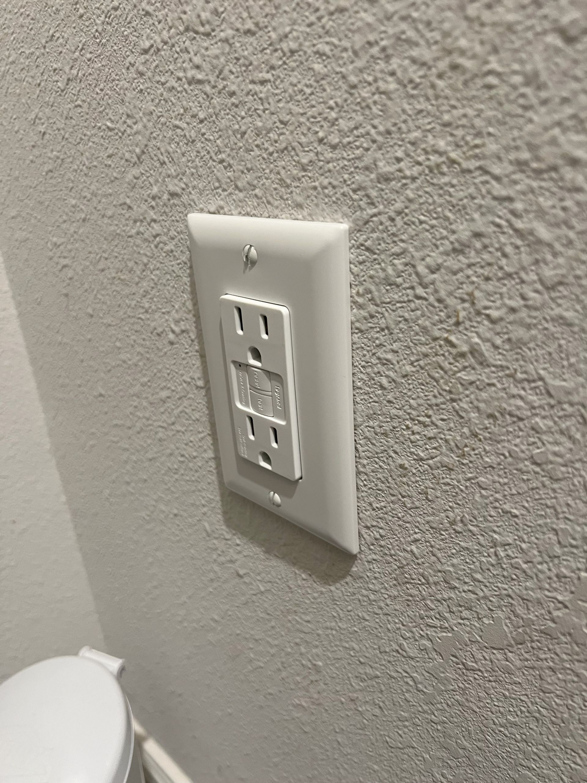 A white electrical outlet on a white wall next to a toilet.