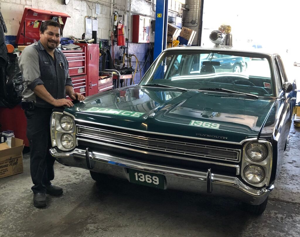 A technician stands beside a dark green vintage car with a prominent chrome grille inside an automotive repair shop. | Hunts Point Service Station