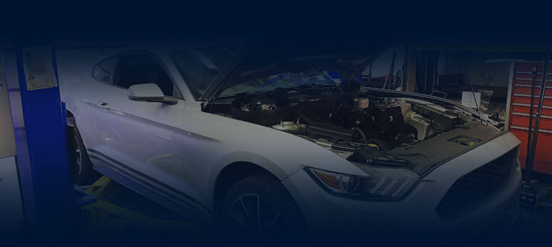 A white Ford Mustang with its hood open, undergoing maintenance inside an auto repair shop with a tool chest nearby. | Hunts Point Service Station