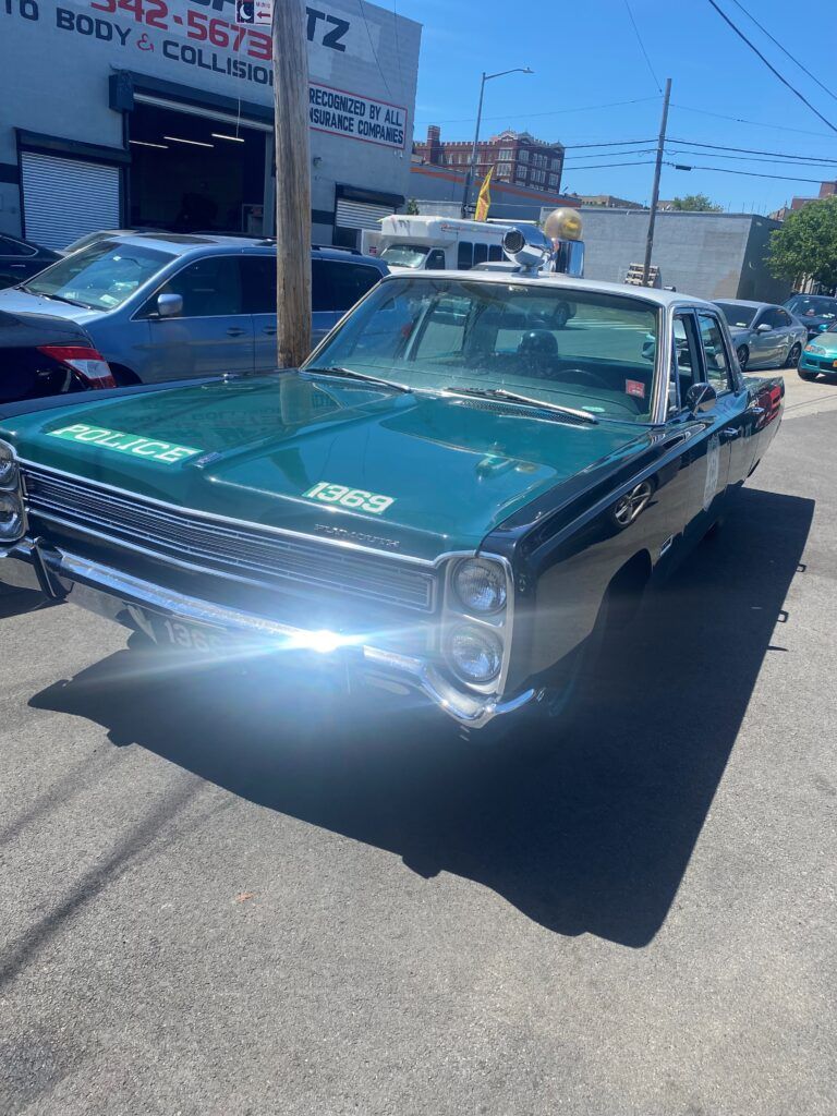 A vintage dark green police car parked on an asphalt street in front of an auto body shop on a sunny day. | Hunts Point Service Station
