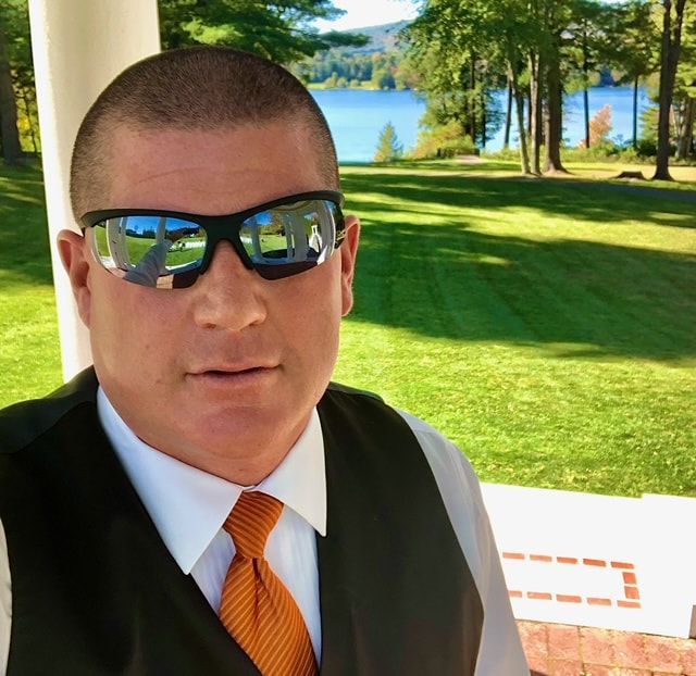 Man in sunglasses, orange tie, and vest, with a lake and trees in the background.