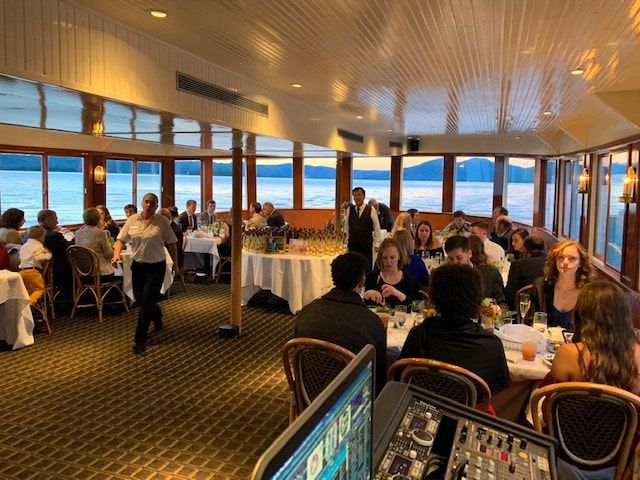 People dining on a boat with large windows overlooking water and mountains, server walking.