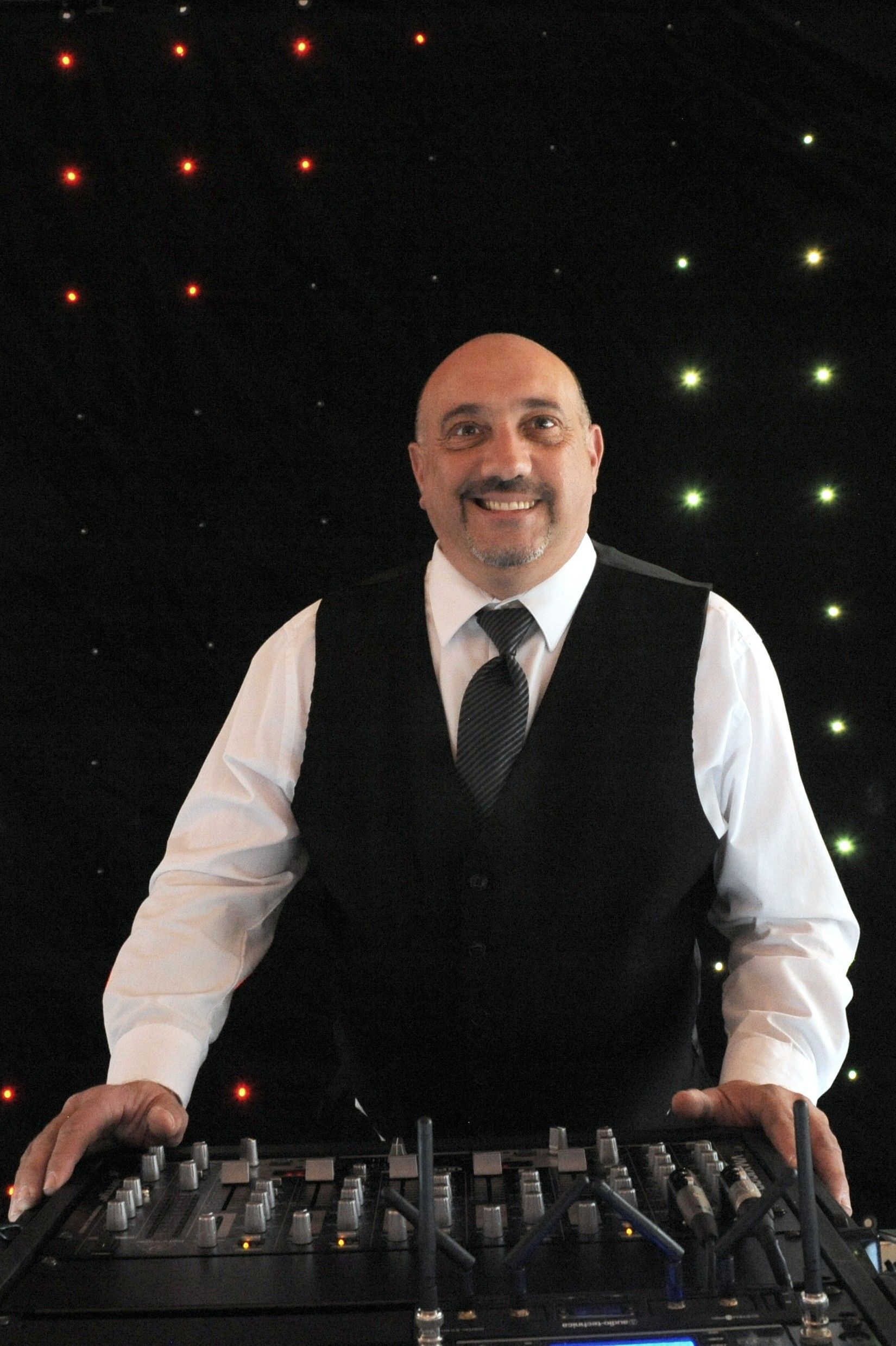 DJ in black vest and tie behind soundboard, smiling, with starry background.
