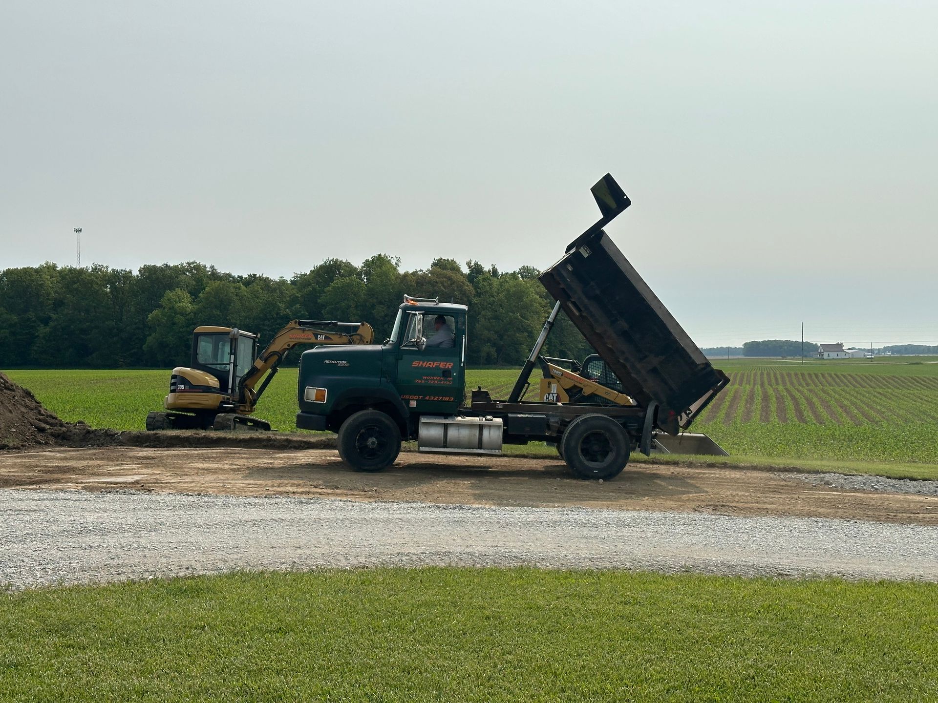 A green dump truck with its bed raised dumps soil next to a small excavator on a construction site.
