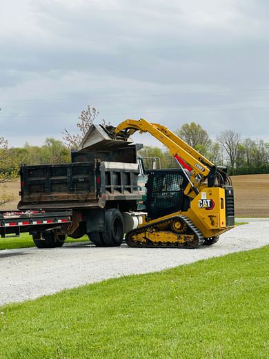 A yellow Caterpillar track loader is dumping dirt into a dump truck on a gravel driveway next to a grassy lawn.