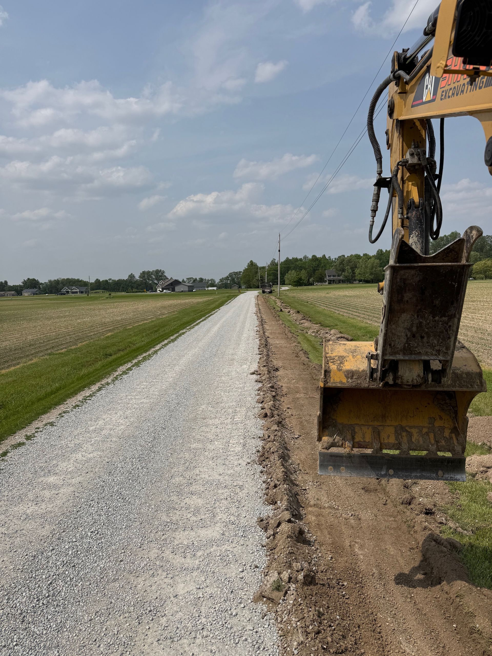 An excavator working on a gravel road.  The road is flanked by green fields and a bright blue sky with puffy clouds.