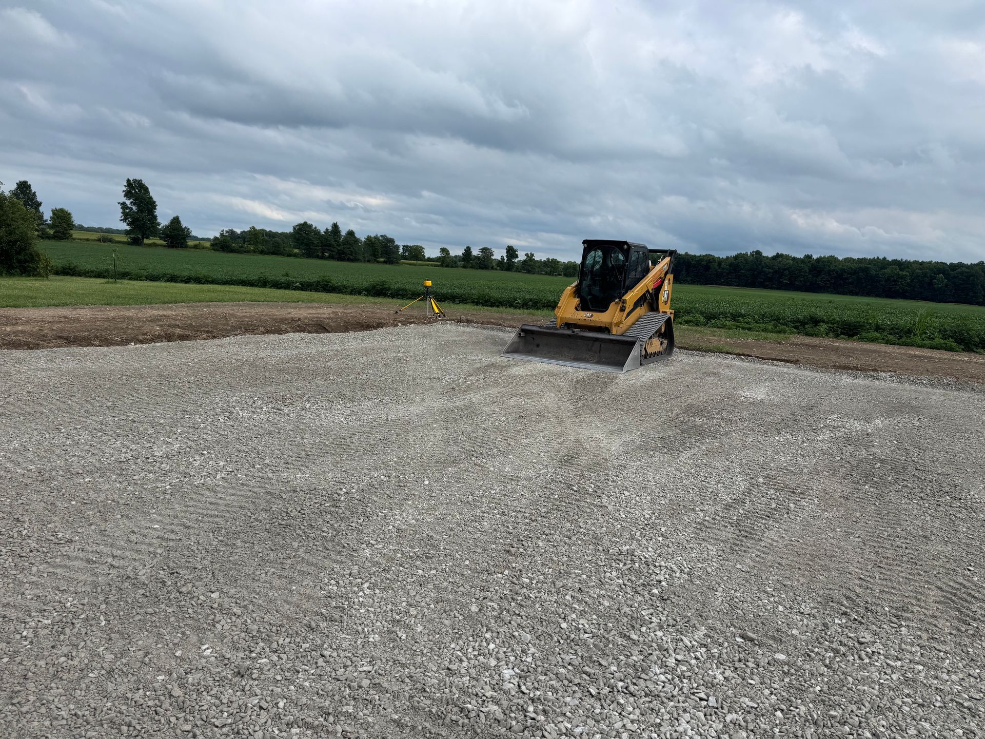 A yellow skid steer loader moving gravel on a construction site under a cloudy sky. Green fields in the background.