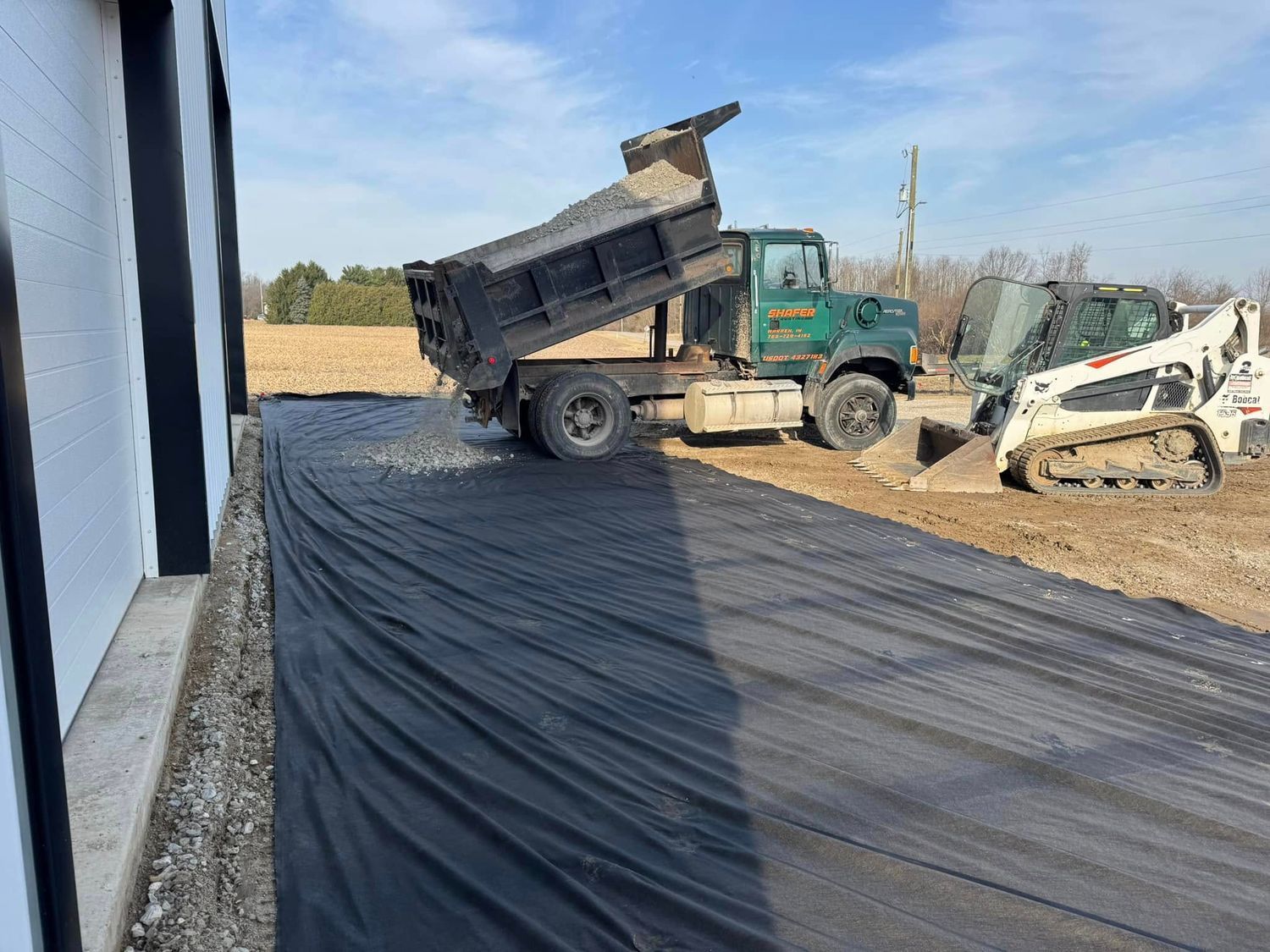 A dump truck unloading gravel onto a black fabric ground cover next to a building. A skid steer sits nearby.