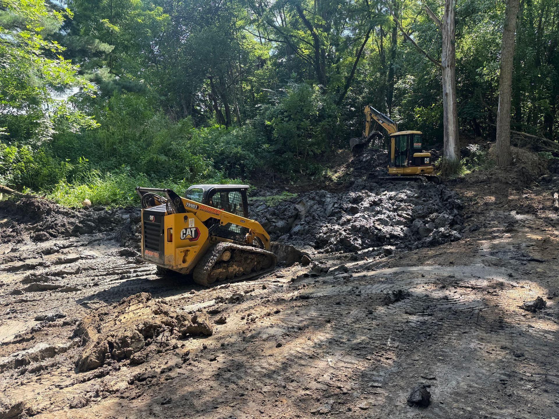 A yellow Caterpillar skid steer and excavator working in a muddy area, clearing land near a tree line.