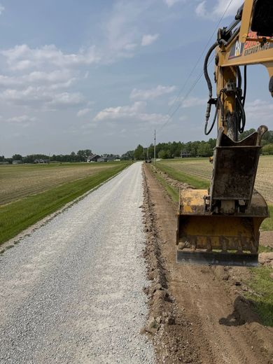 A gravel road being graded by a yellow excavator. Fields and a bright sky are in the background.