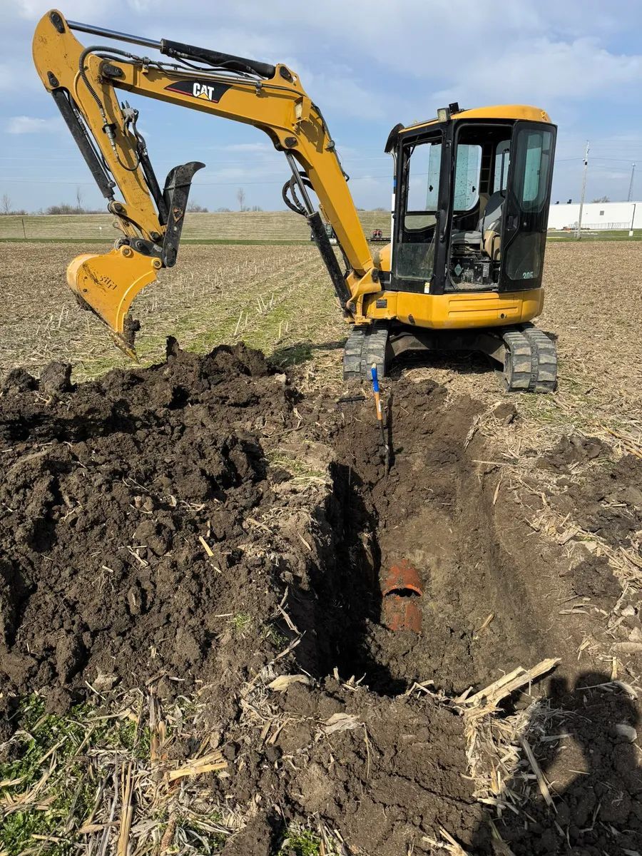Yellow excavator digging a trench in a field. A rusty metal object is visible in the trench.