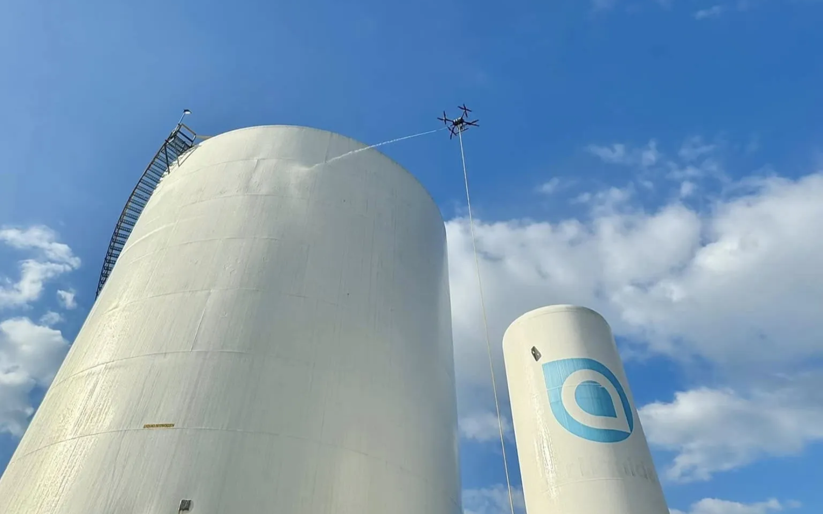 Drone flying, stringing a cable from a high white silo under a blue sky with clouds.