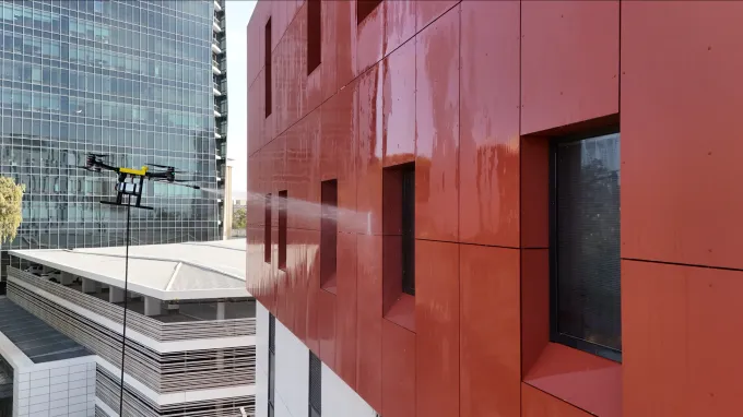 Drone spraying water on an orange building facade, near another glass skyscraper.