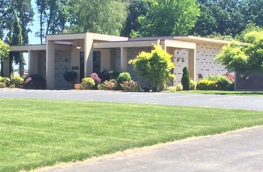 Tan building with multiple open sections and a green lawn. Trees and bushes surround.