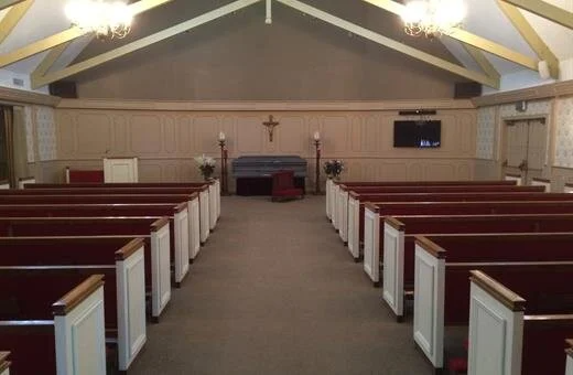 Interior of a chapel with rows of red-cushioned pews facing an elevated coffin, cross, and flowers.