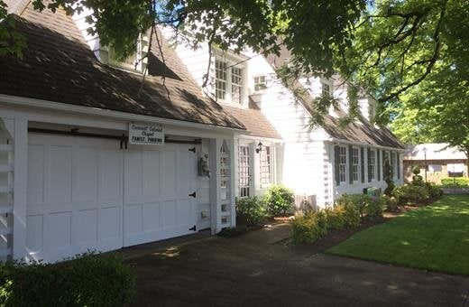 White building with a garage, windows, and a manicured lawn under the shade of trees.