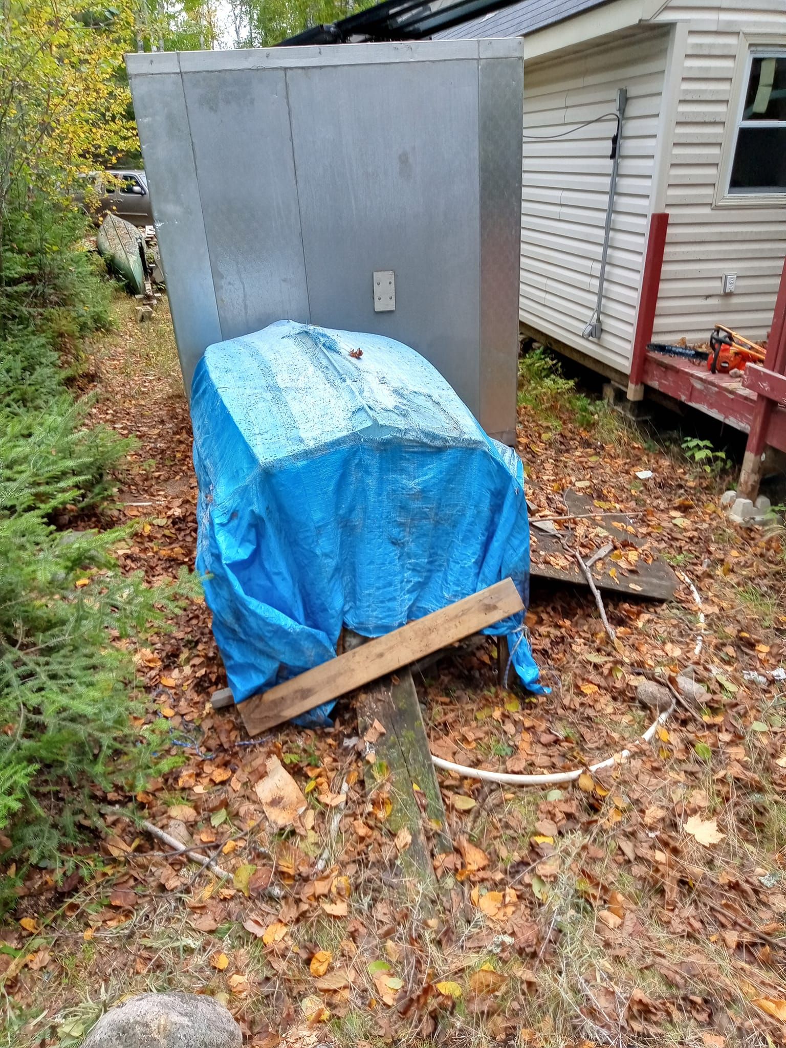 Blue tarp covers something on a wood support in a yard, with a metal box and house in the background.