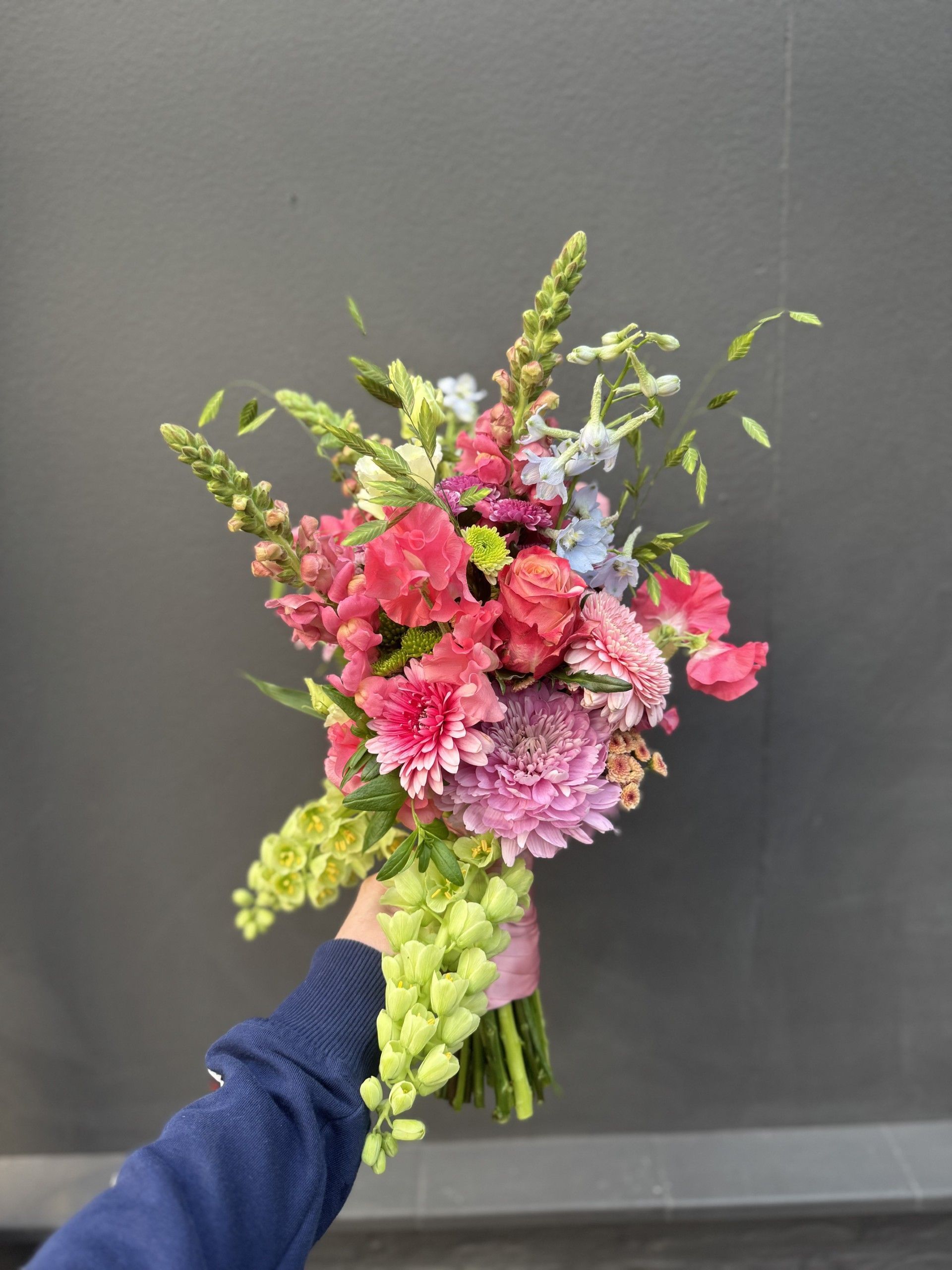 Een hand houdt een levendig boeket met roze, lichtpaarse, lichtblauwe en limoengroene bloemen vast tegen een effen grijze muur.