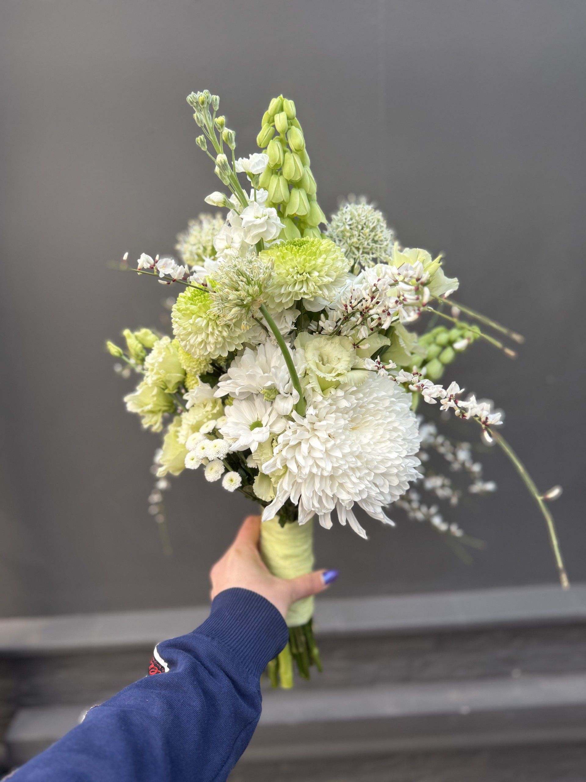 Een persoon houdt een bruidsboeket met witte en lichtgroene bloemen, waaronder chrysanten, tegen een grijze muur.
