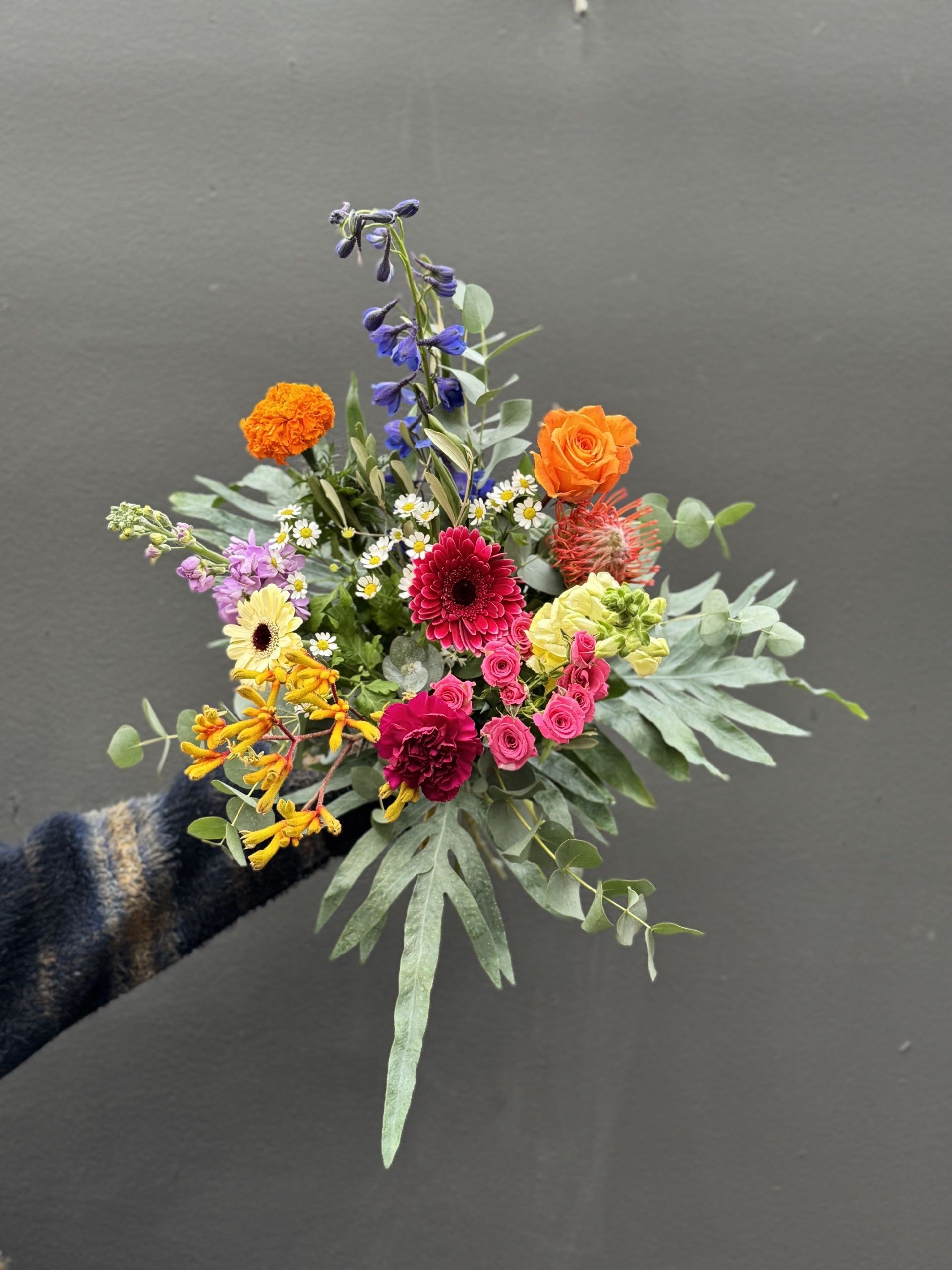 Een persoon houdt een levendig boeket vast met oranje, roze, gele en blauwe bloemen en groene eucalyptusbladeren.