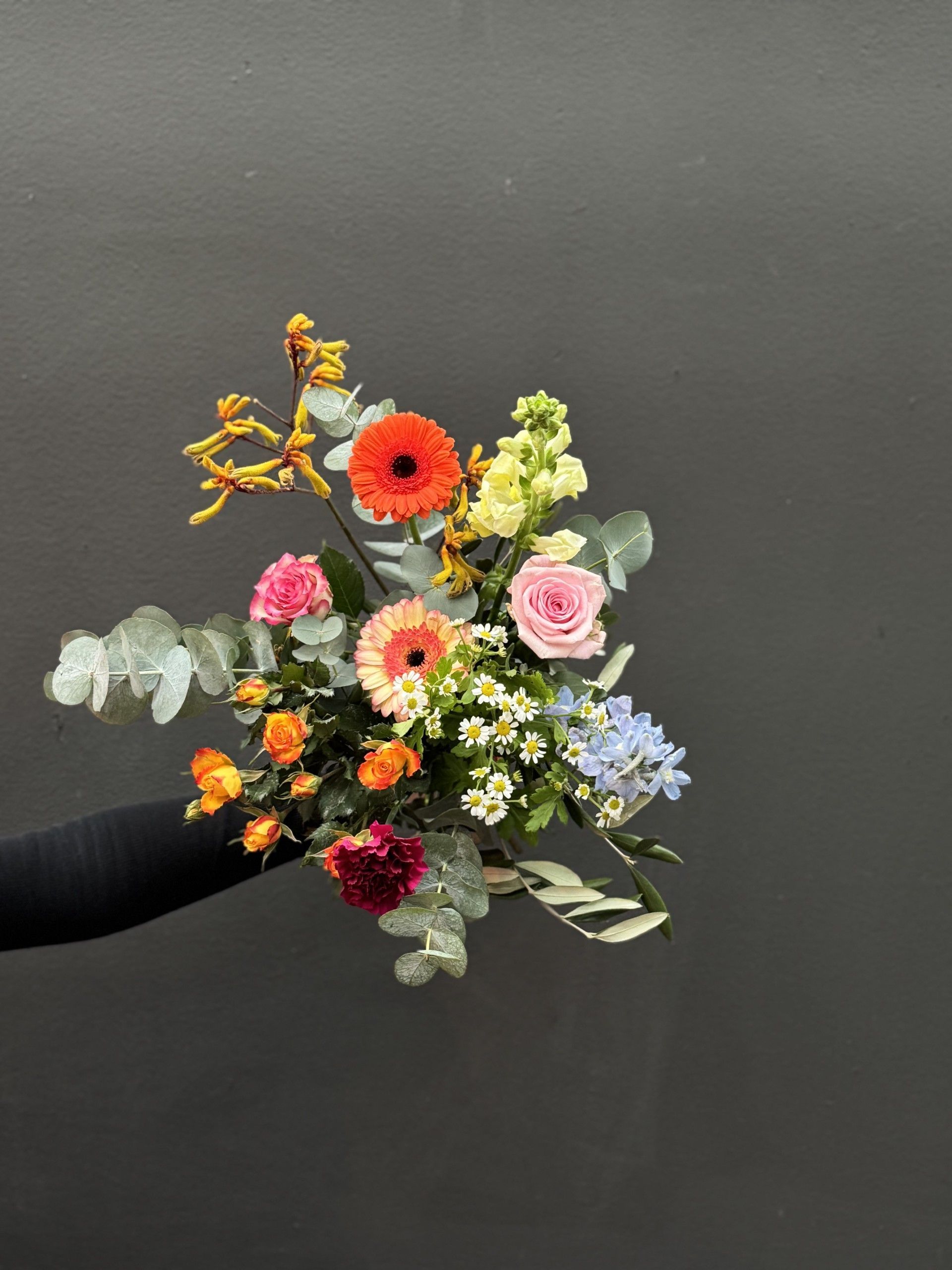 Een kleurrijk boeket met een oranje gerbera, roze rozen, gele leeuwenbekjes en eucalyptus, tegen een muur gehouden.