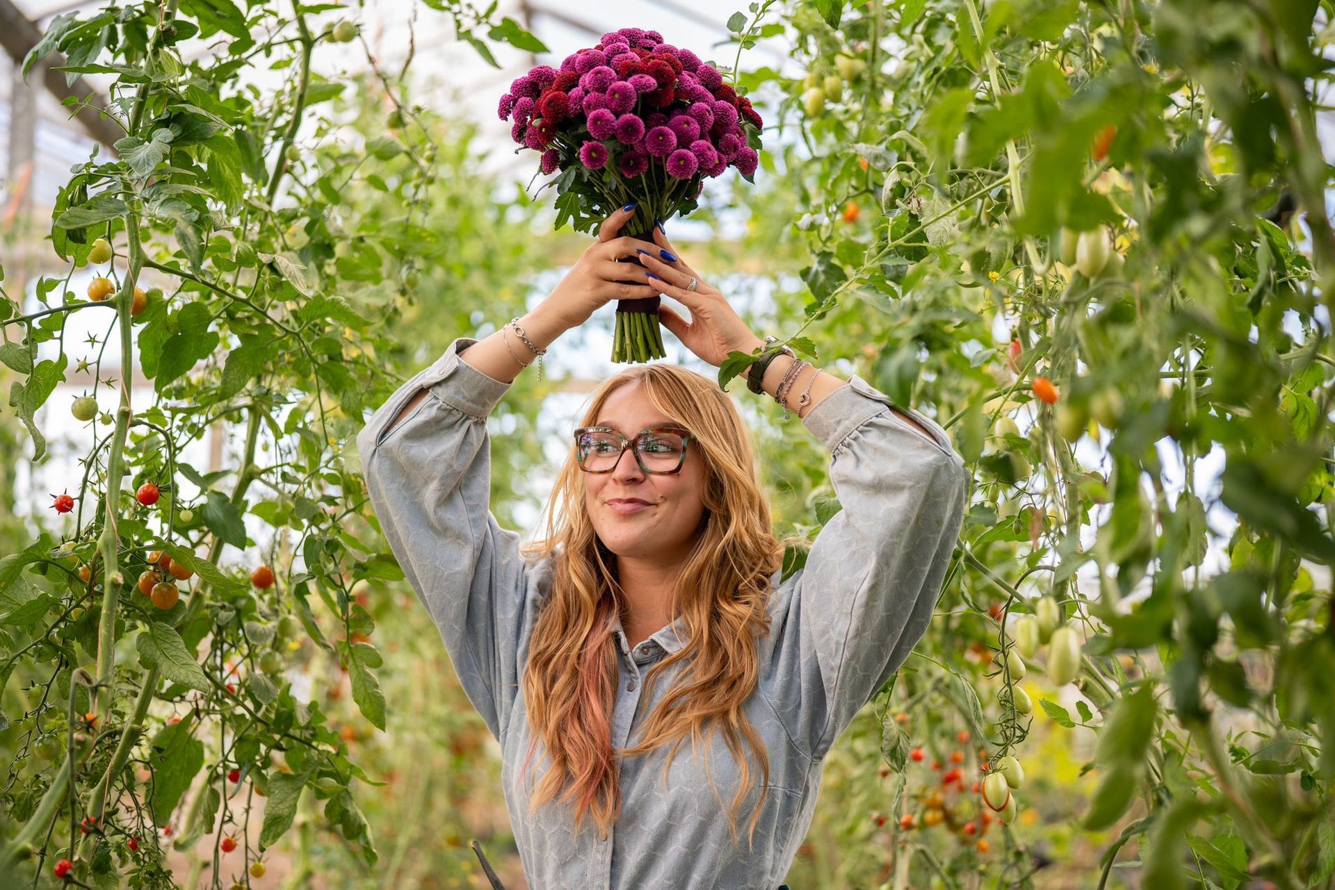 Een glimlachend persoon met een bril staat in een kas en houdt een boeket dieppaarse bloemen boven het hoofd.