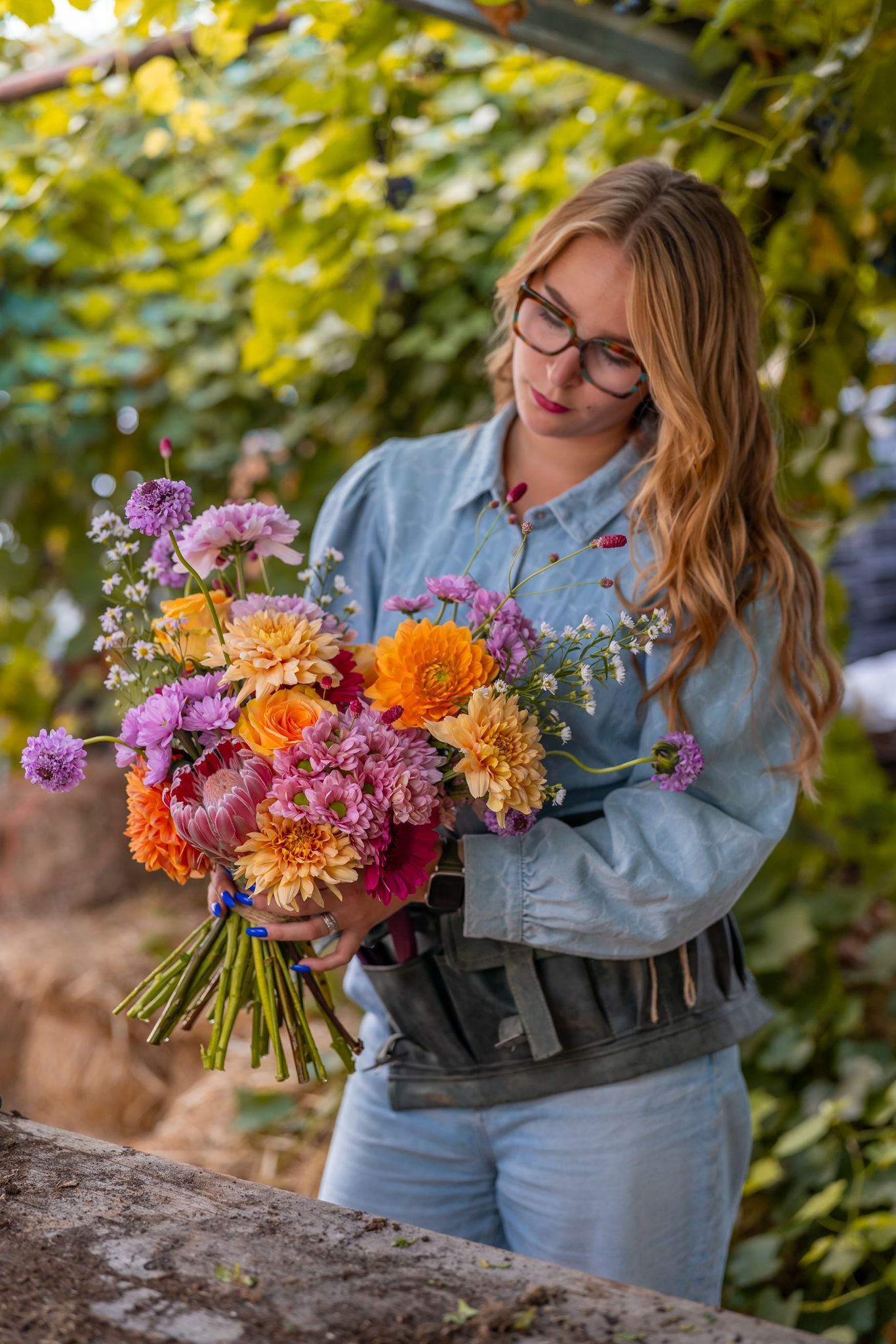 Een persoon in een spijkerhemd houdt een levendig boeket oranje, roze en paarse bloemen vast in een weelderige buitentuin.