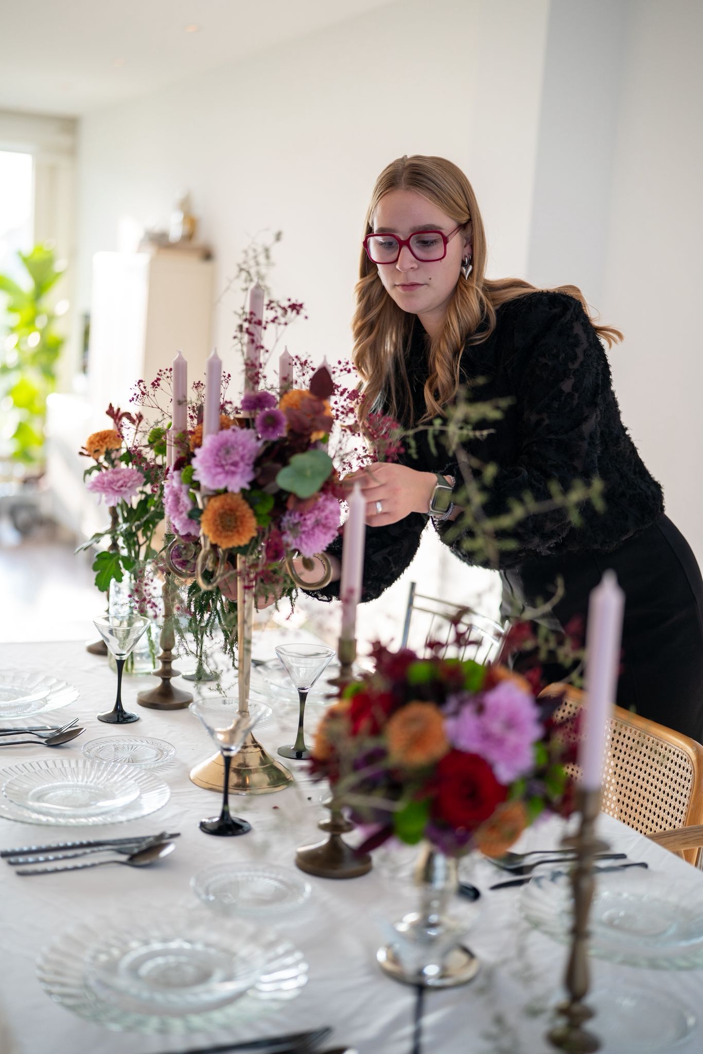 Een persoon in een zwart shirt schikt bloemstukken met kaarsen op een tafel die gedekt is voor een formeel diner.