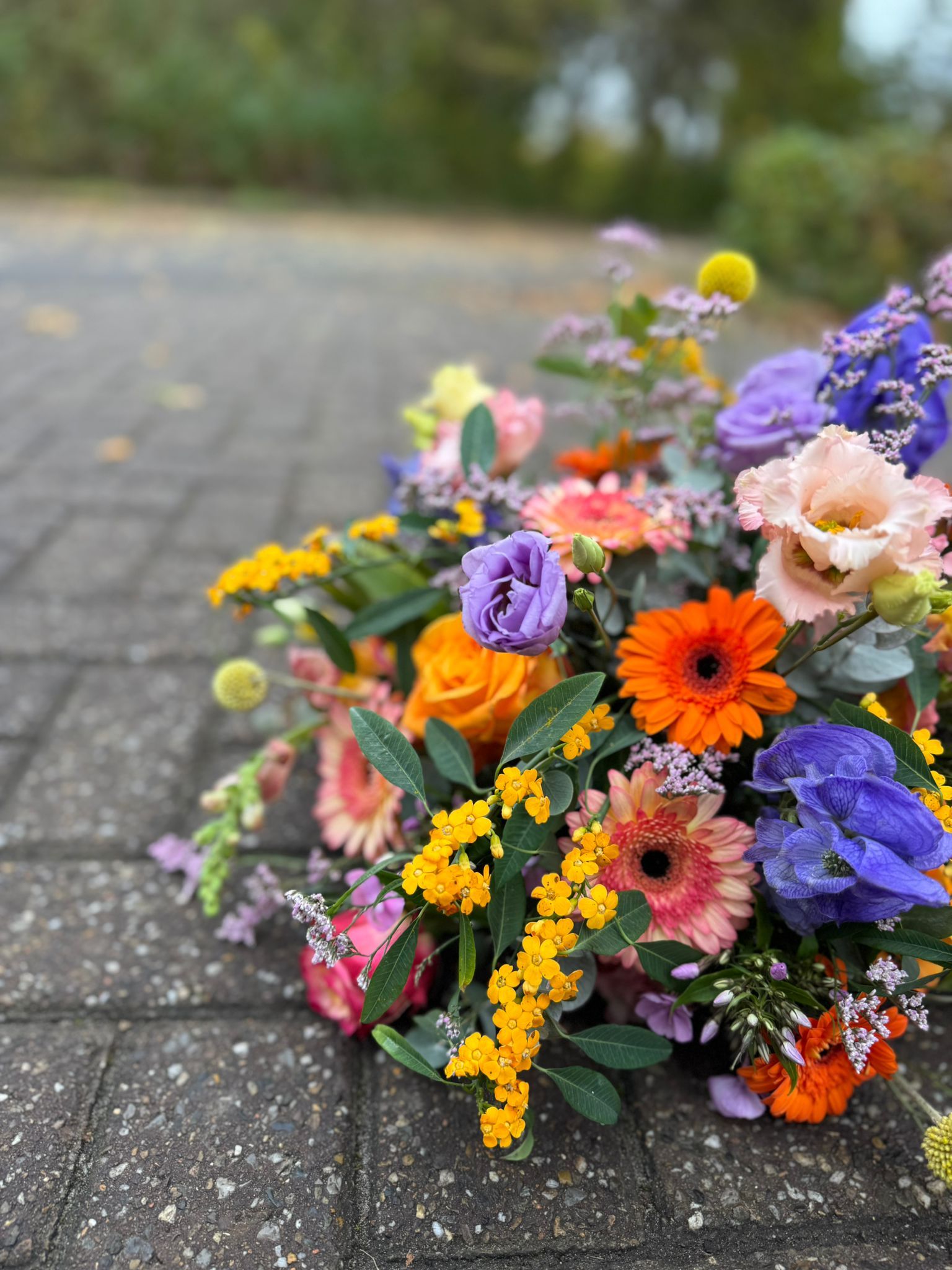 Een levendig, kleurrijk bloemenboeket met oranje gerbera's, paarse lisianthus en gele solidago op een stenen pad.