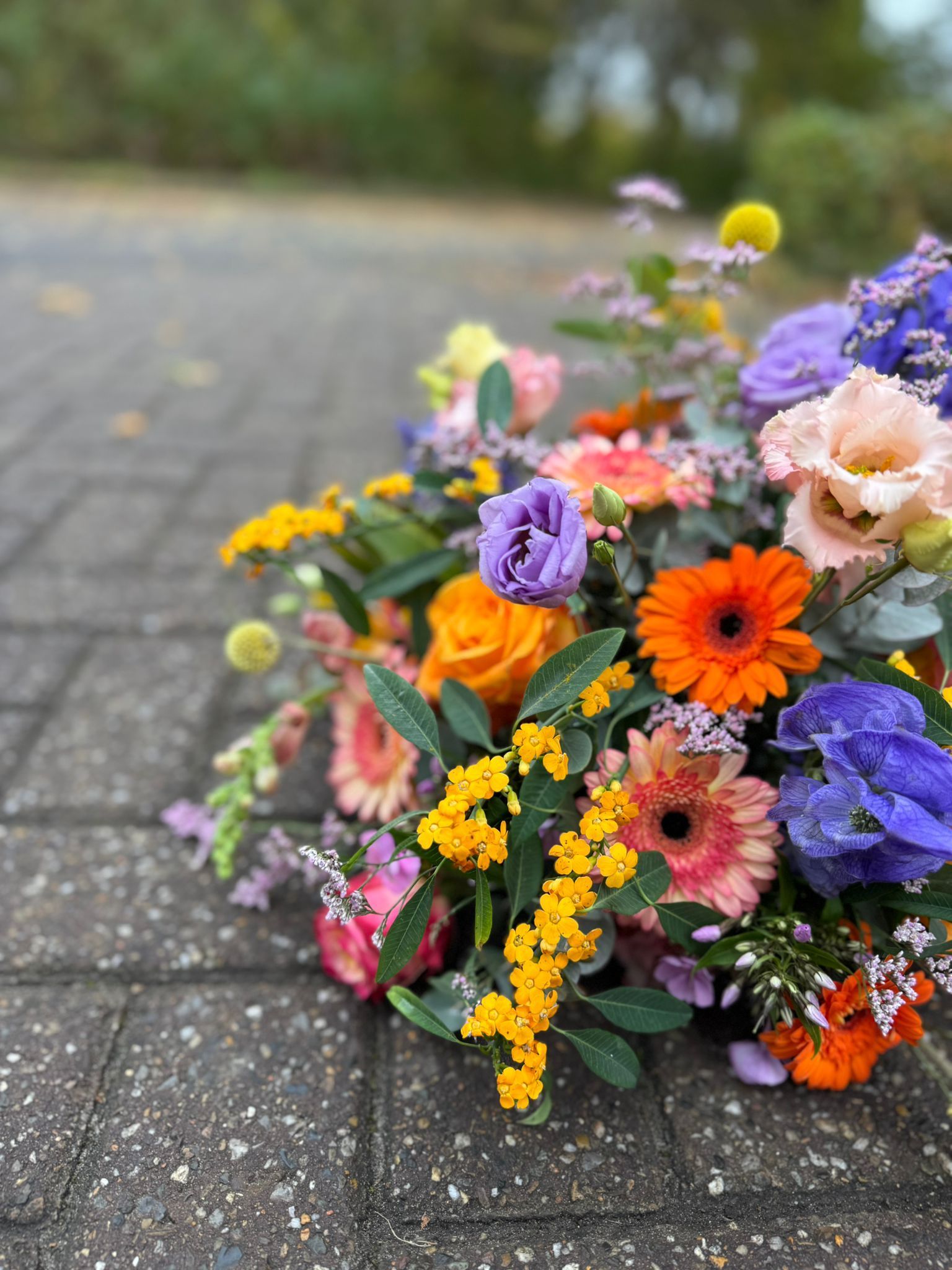 Een levendig, kleurrijk bloemenboeket met oranje gerbera's, paarse lisianthus en rozen op een stenen pad.