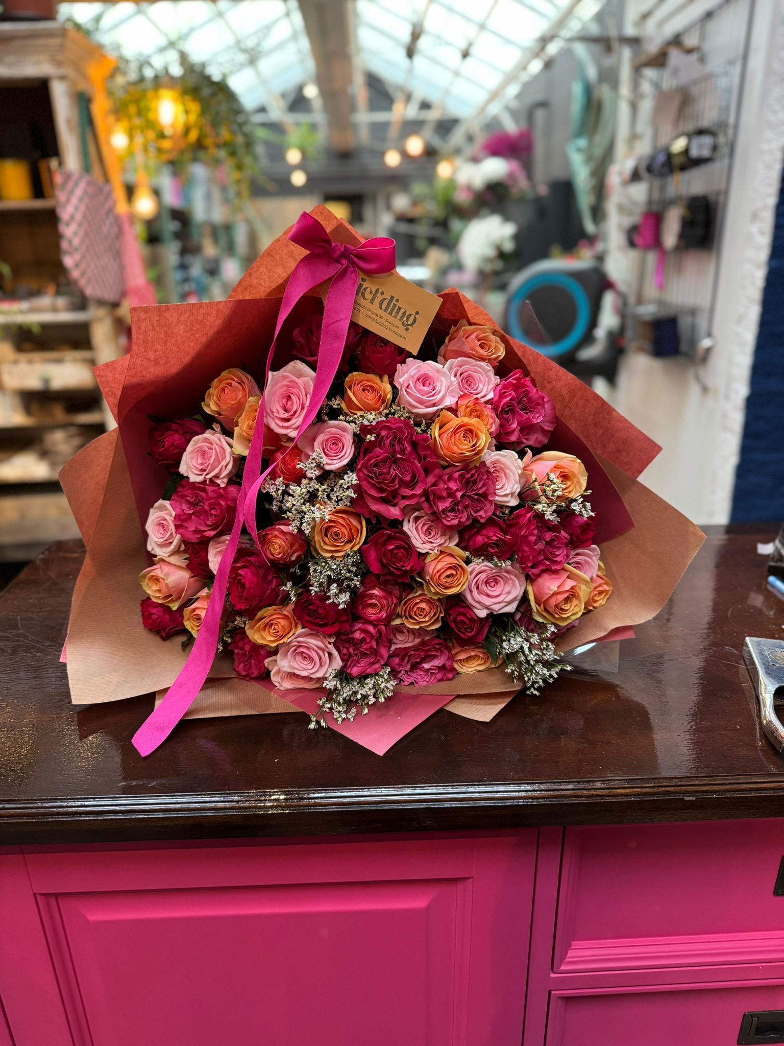 Een levendig boeket roze, oranje en rode bloemen, verpakt in bruin papier met een felroze strik, staat op een toonbank.
