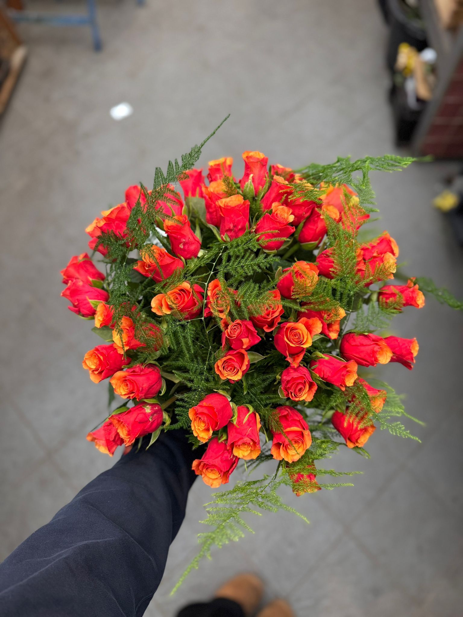 Een boeket oranje en gele rozen met groen blad, in iemands hand gehouden tegen een grijze vloerachtergrond.