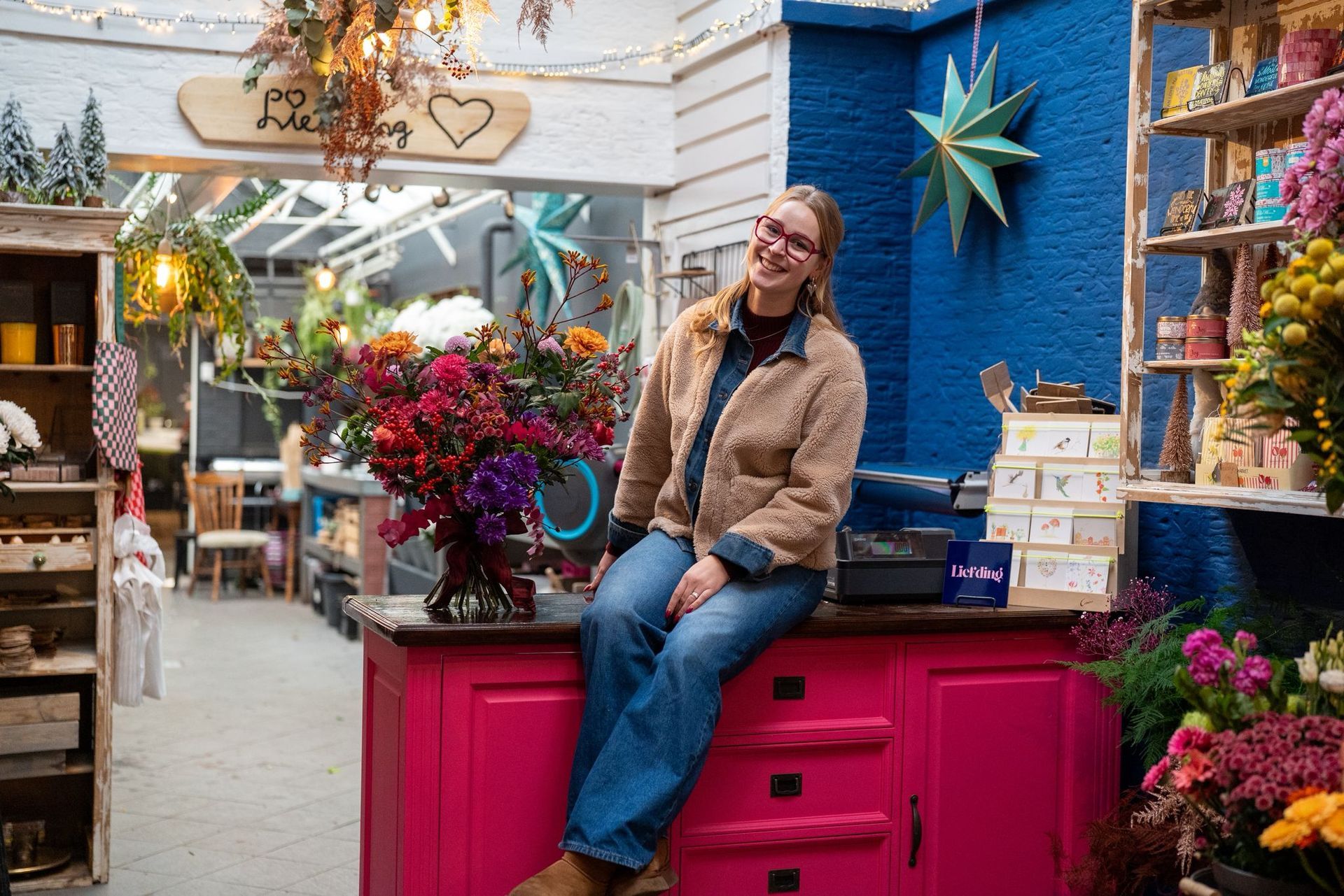 Een persoon in een beige fleecevest zit op een felroze toonbank in een bloemenwinkel, naast een groot boeket tegen een blauwe muur.