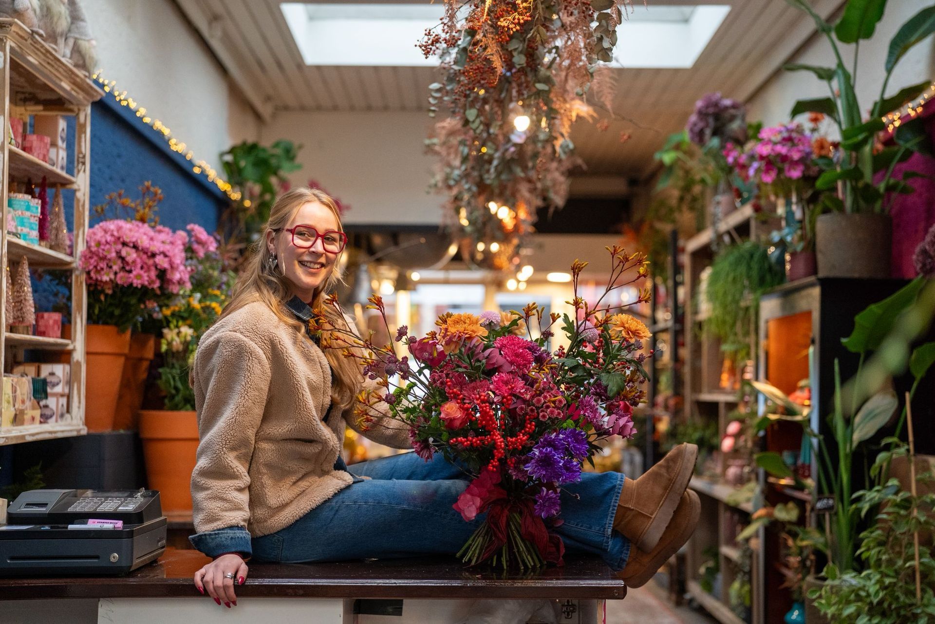 Een bloemist zit glimlachend op een houten toonbank in een bloemenwinkel, terwijl hij een groot, kleurrijk boeket bloemen vasthoudt.