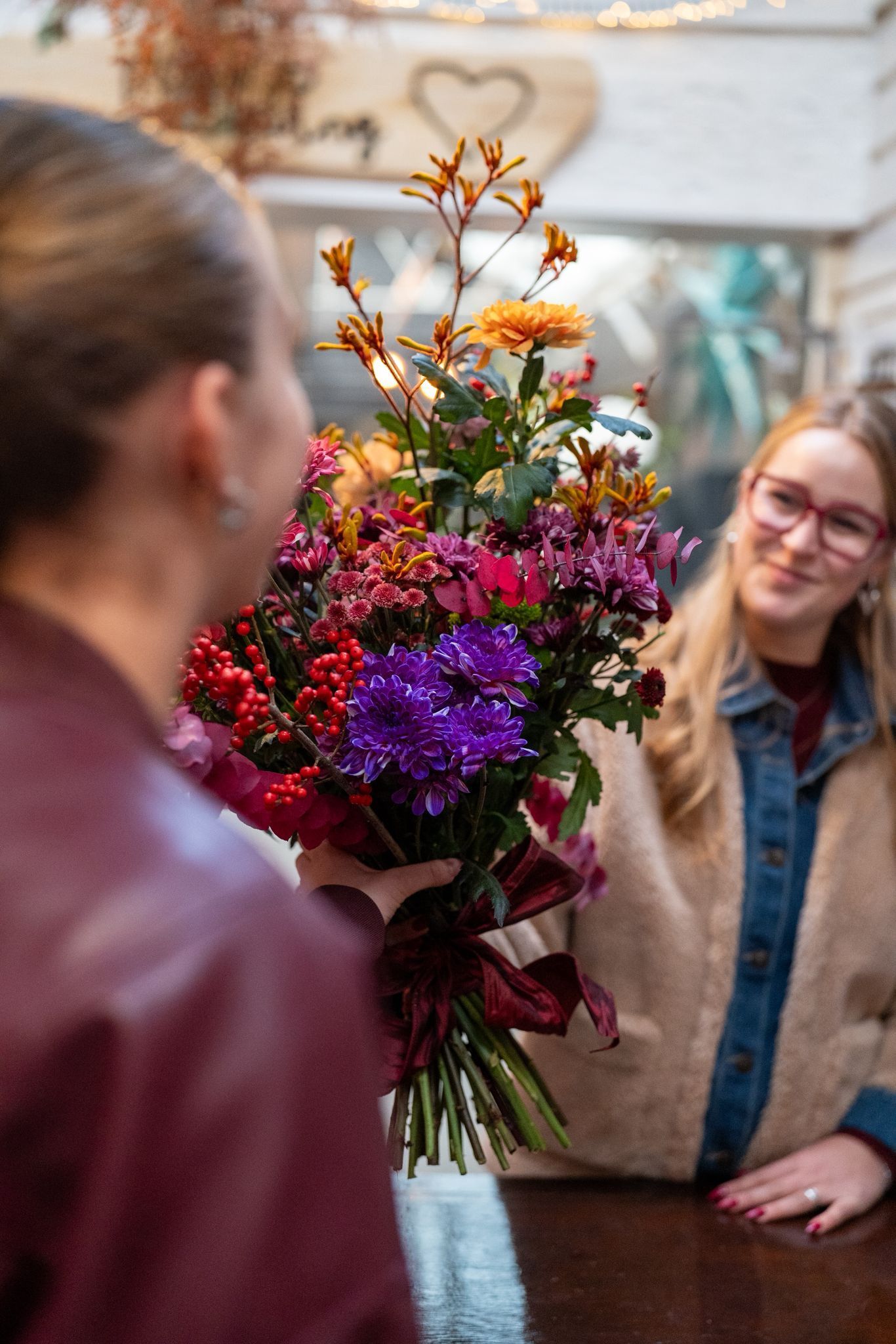 Iemand overhandigt een levendig, veelkleurig boeket bloemen aan een ontvanger die achter een houten toonbank staat.