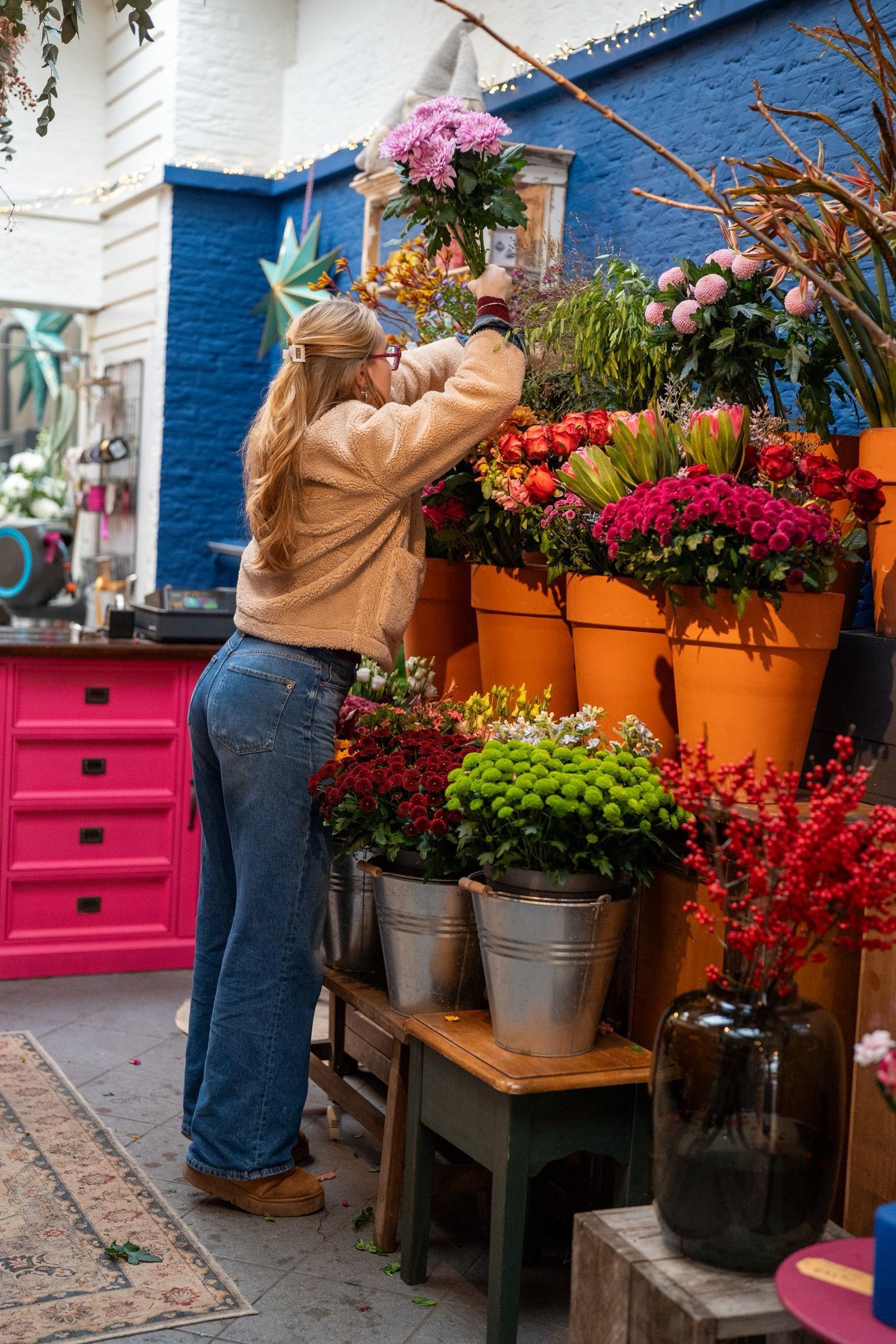 Een persoon in een beige fleecejack en blauwe spijkerbroek schikt een boeket bloemen in een winkel met feloranje plantenbakken.
