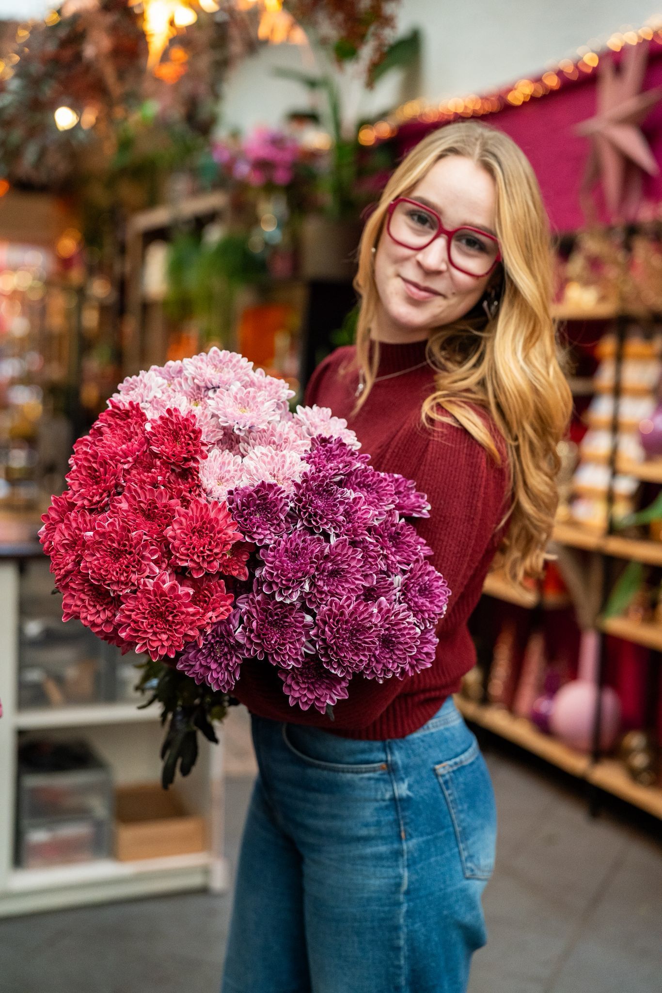 Een lachende persoon met een groot, kleurrijk boeket roze, rode en paarse anjers in een lichte bloemenwinkel.